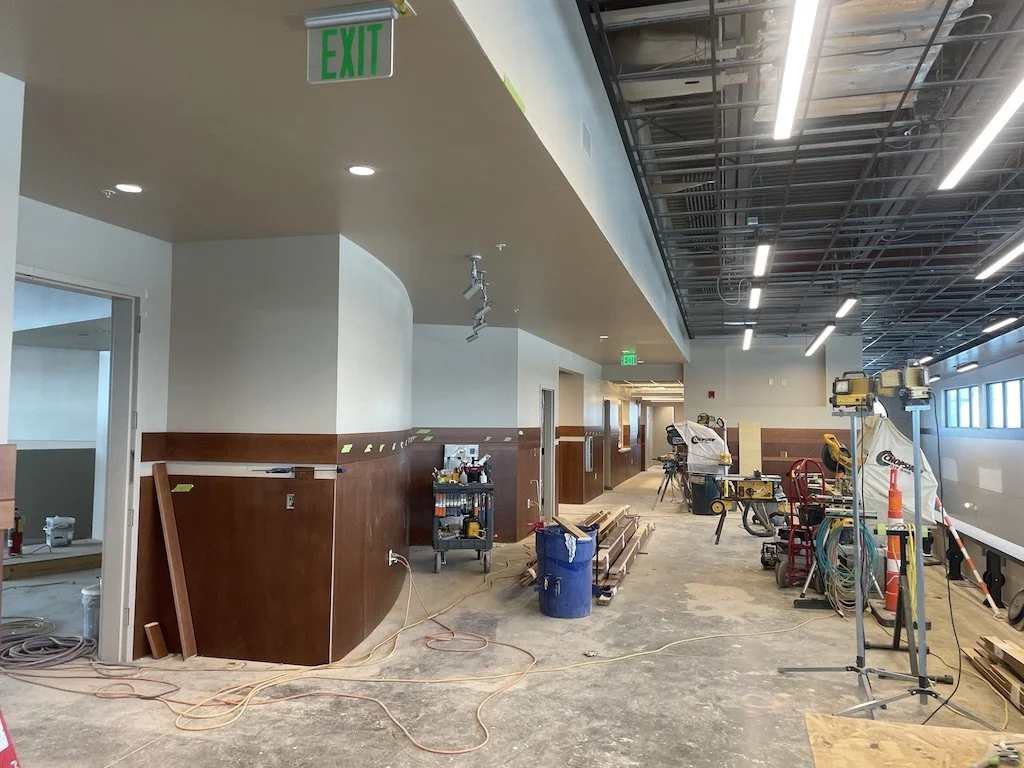 Interior of a building under construction or renovation, showing unfinished floors, construction tools, equipment, and materials scattered around, with a wall featuring wood paneling, and an EXIT sign hanging from the ceiling.