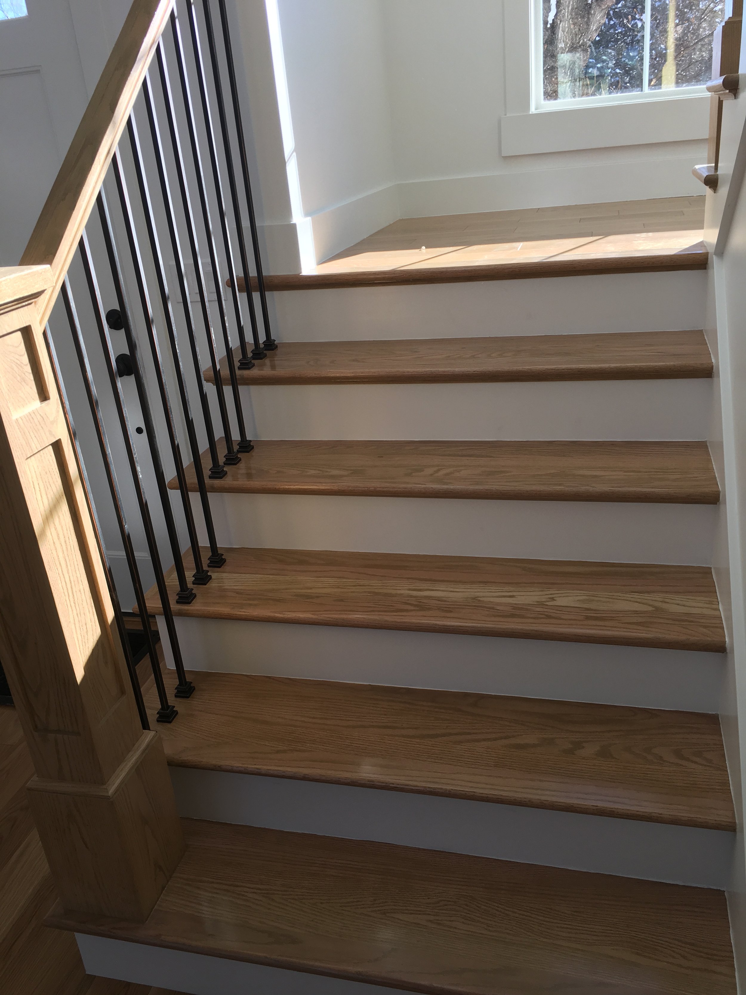 Interior view of a staircase with wooden steps and a black metal railing, adjacent to a white wall and a large window showing outdoor trees and sky.