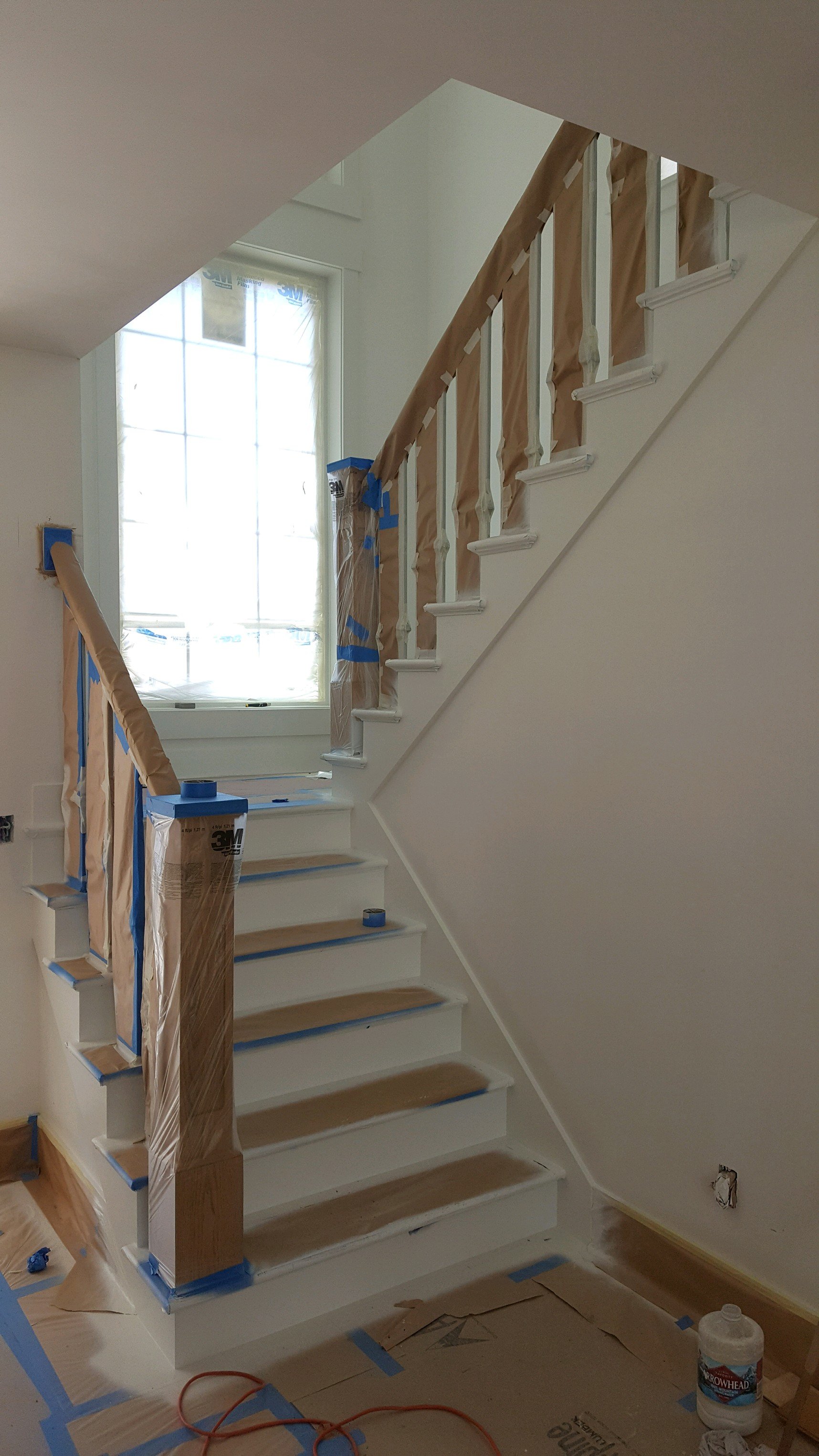 Interior of a staircase under renovation, with protective paper covering the handrail and new white paint on the stairs, taped off for painting or finishing work.