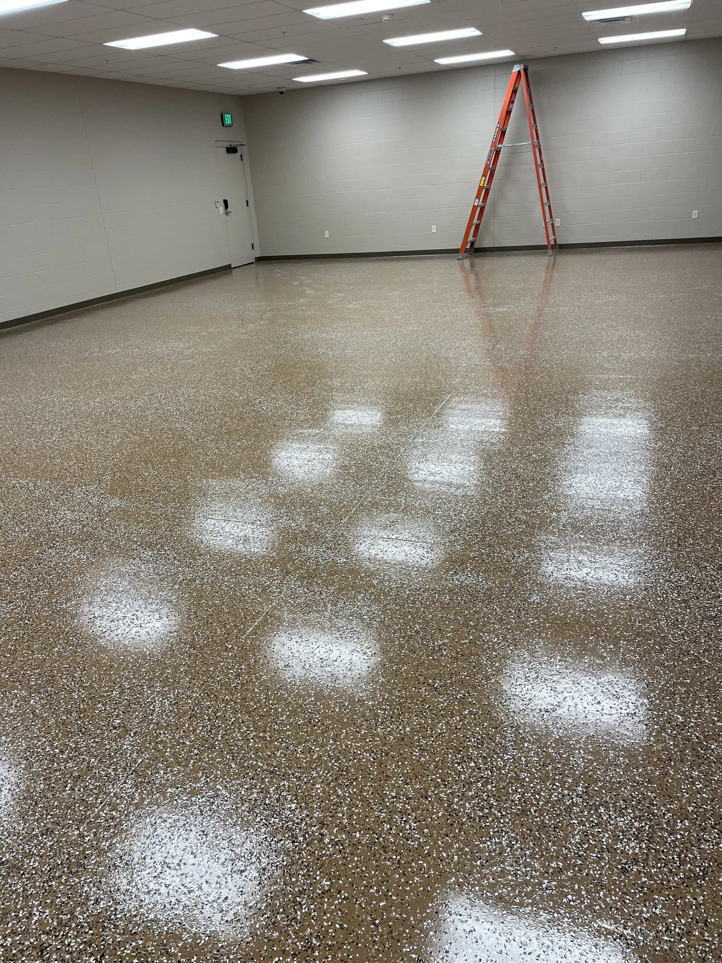 Empty room with beige speckled flooring, white walls, fluorescent ceiling lights, a closed door, and an orange ladder leaning against the wall.