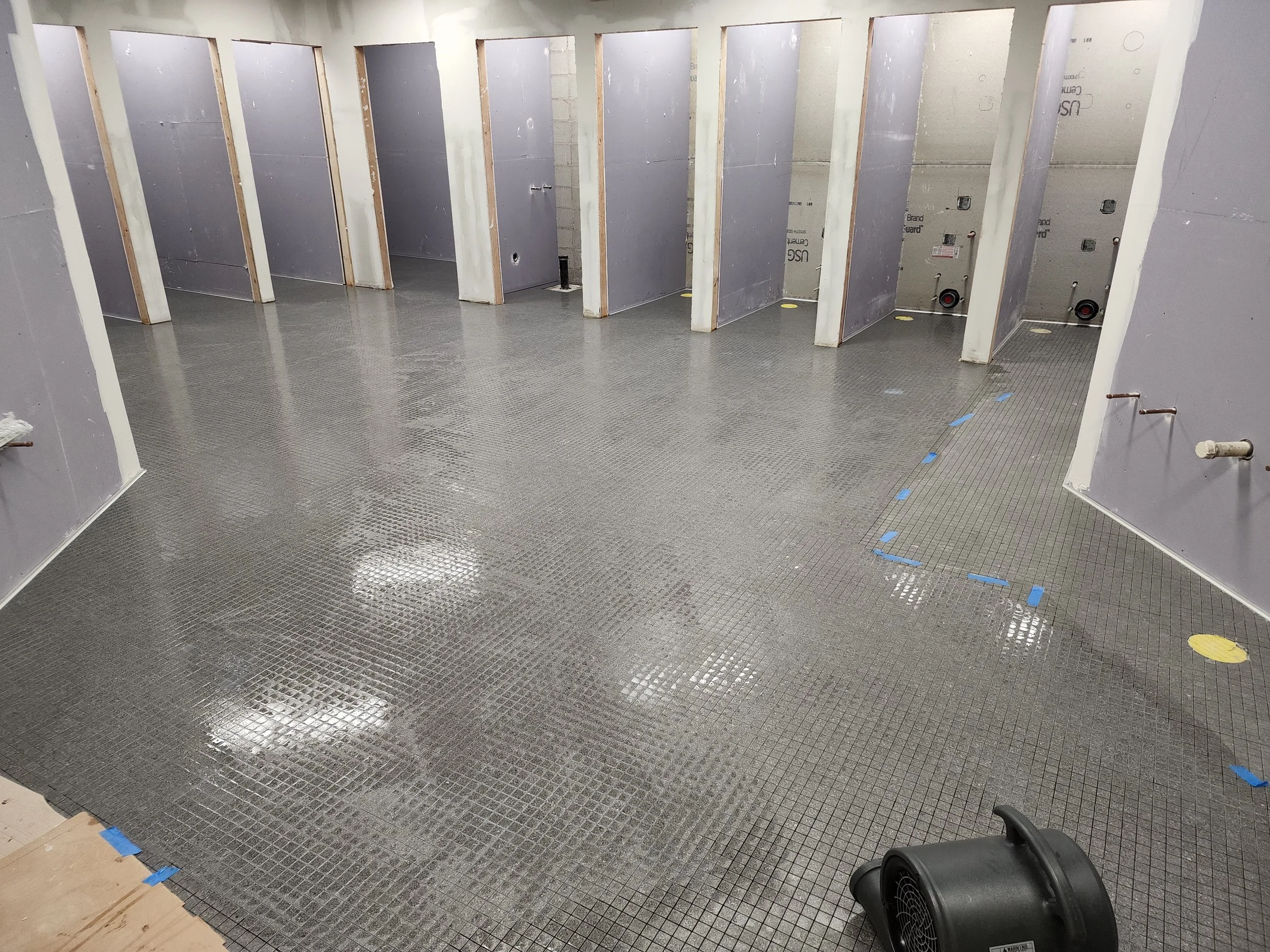 Interior of a building under construction with drywall partitions, exposed wiring and plumbing, and black flooring.