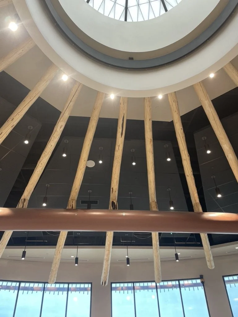 Interior view of a building with a circular skylight ceiling, wooden beams hanging down, and modern lighting fixtures.