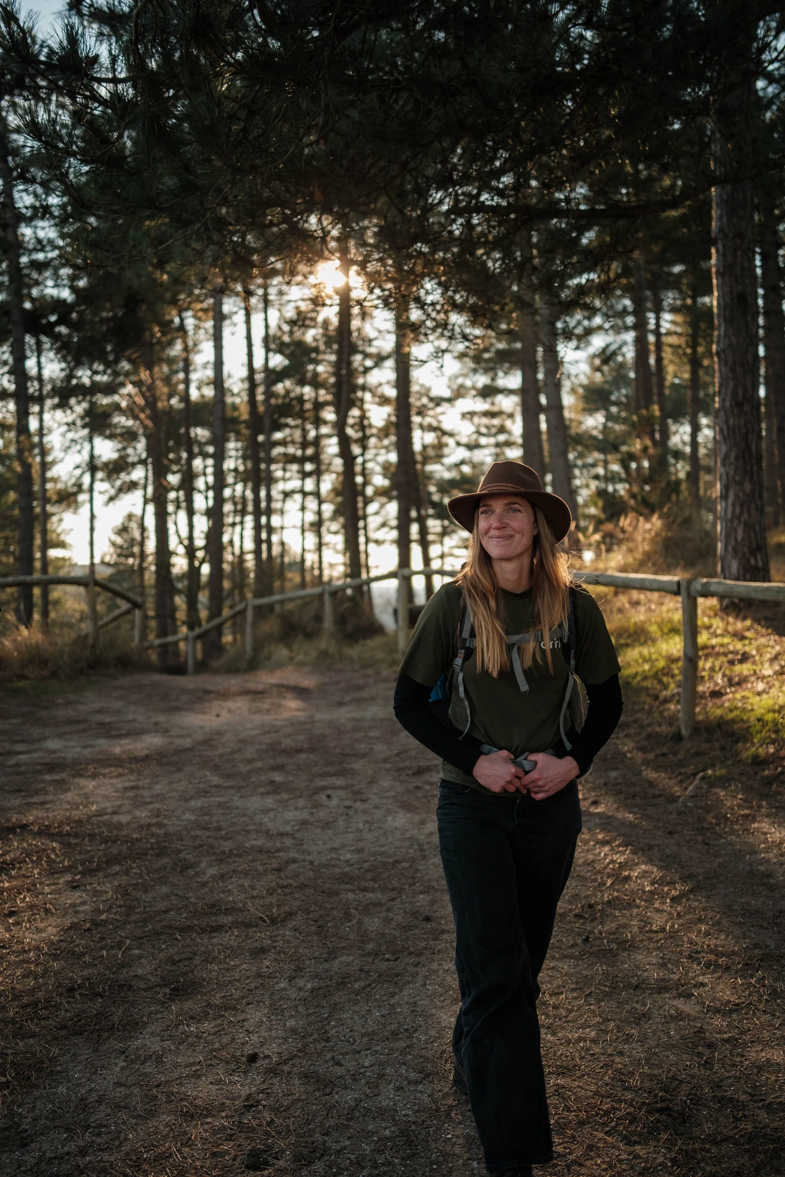 Vrouw wandelt in het bos tijdens zonsondergang, draagt hoed en backpack, glimlacht.