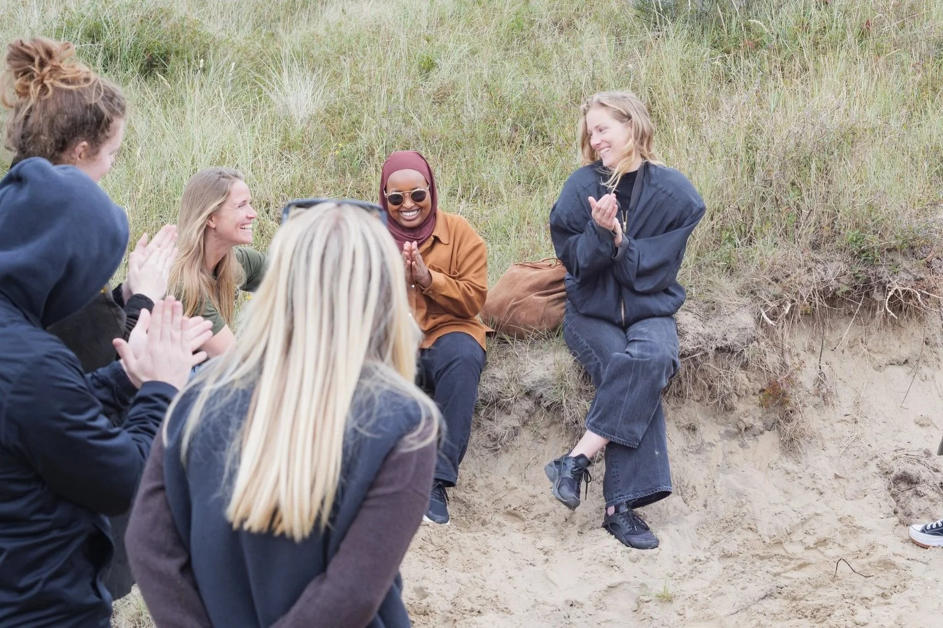 Vrouw zit op een zandheuvel terwijl een groep mensen haar applaudisseert en lacht.