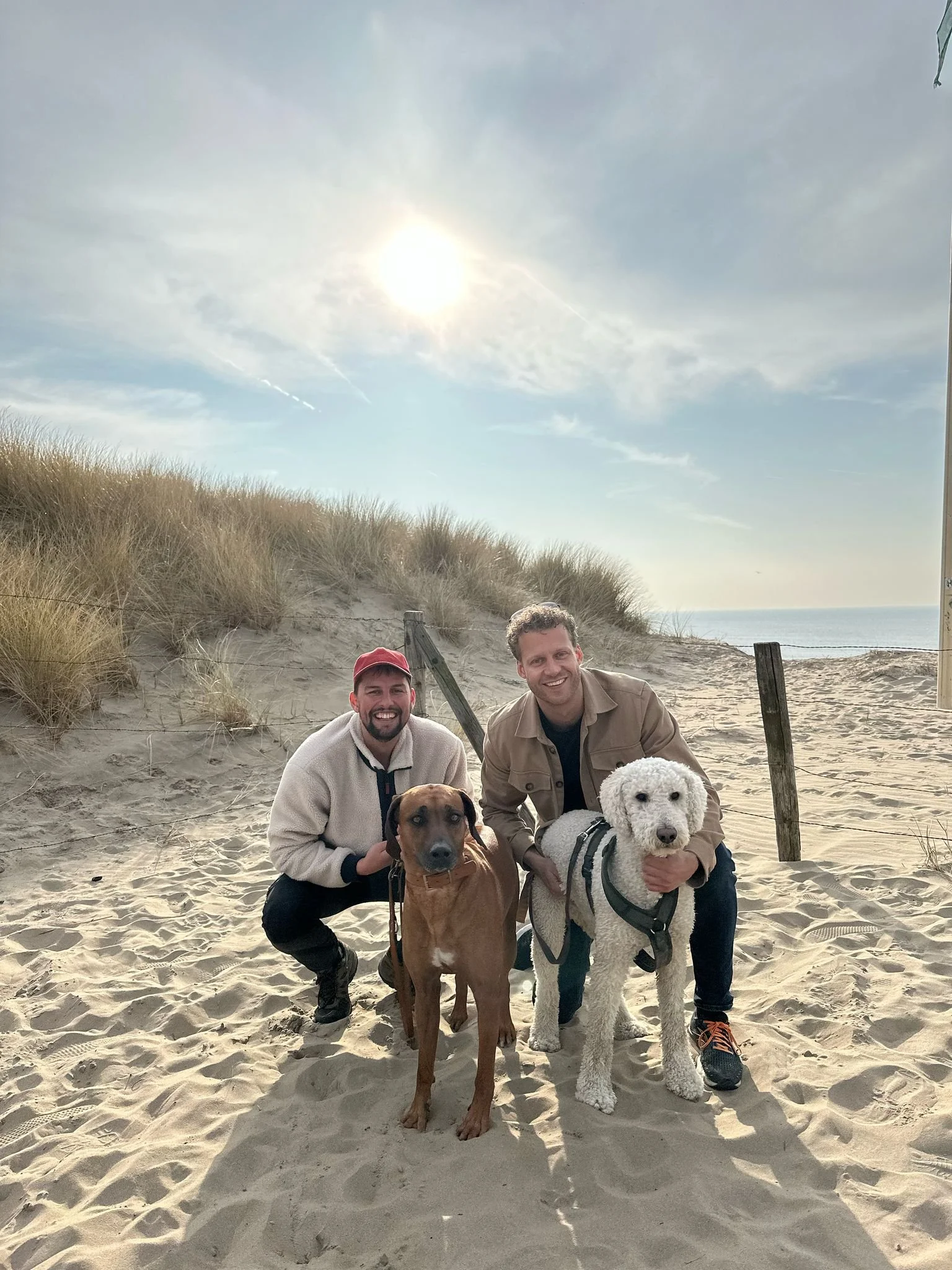 Twee mannen poseren op het strand met twee honden, onder een heldere zon, omgeven door zand en duinen.