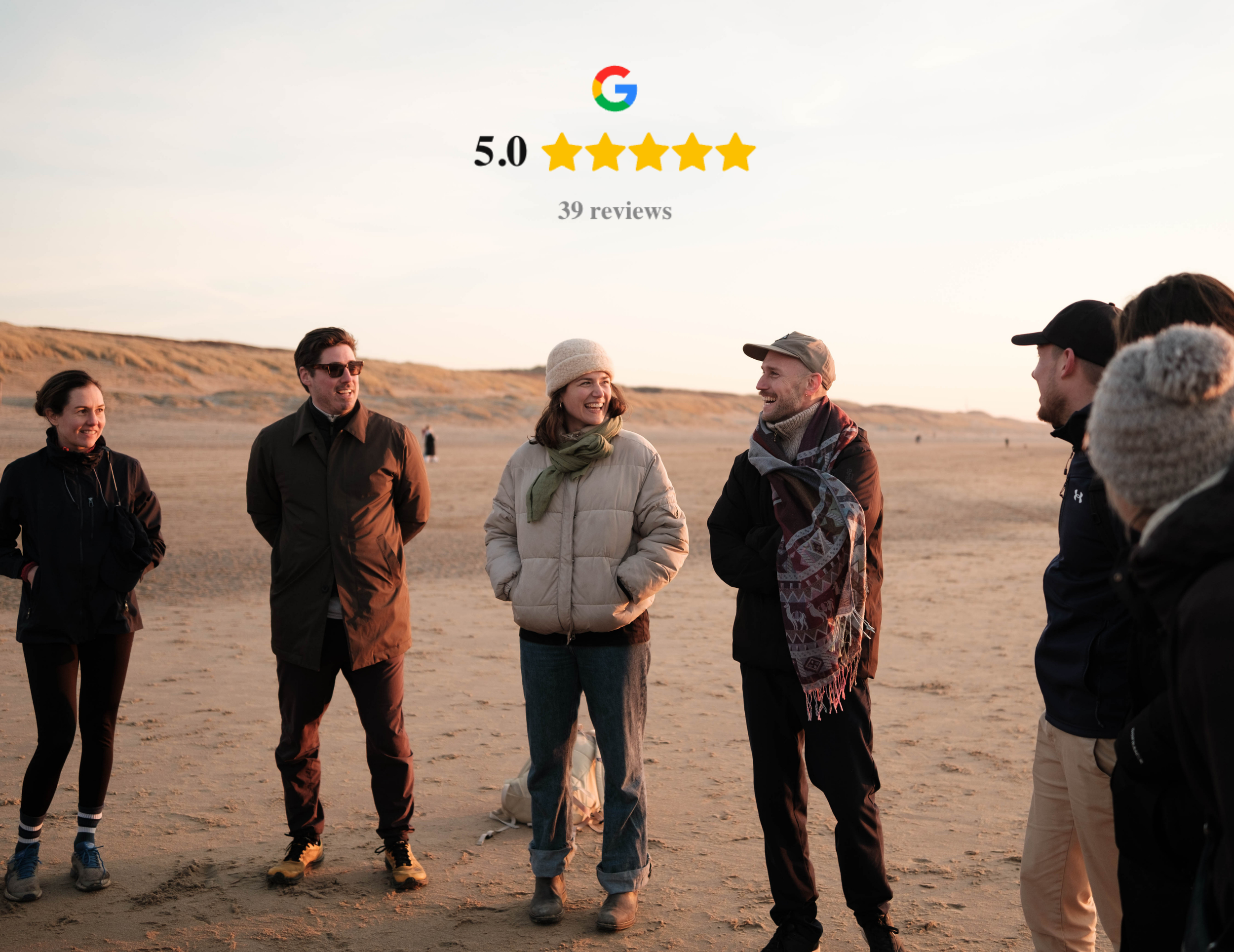 Een groep mensen die buiten op een zandstrand lopen, lachend en praten, met een ondergaande zon en duinen op de achtergrond.