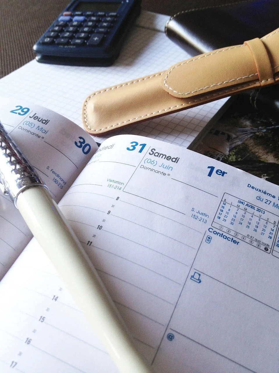 Open planner or diary showing dates March 29 to April 2, 2013, with a pen resting on it. Items on the desk include a calculator, a beige leather strap, a flashlight, a black object, and a sheet of grid paper.