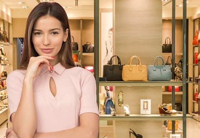 Woman with long brown hair in a pink blouse shopping in a handbags store with shelves displaying handbags in the background.