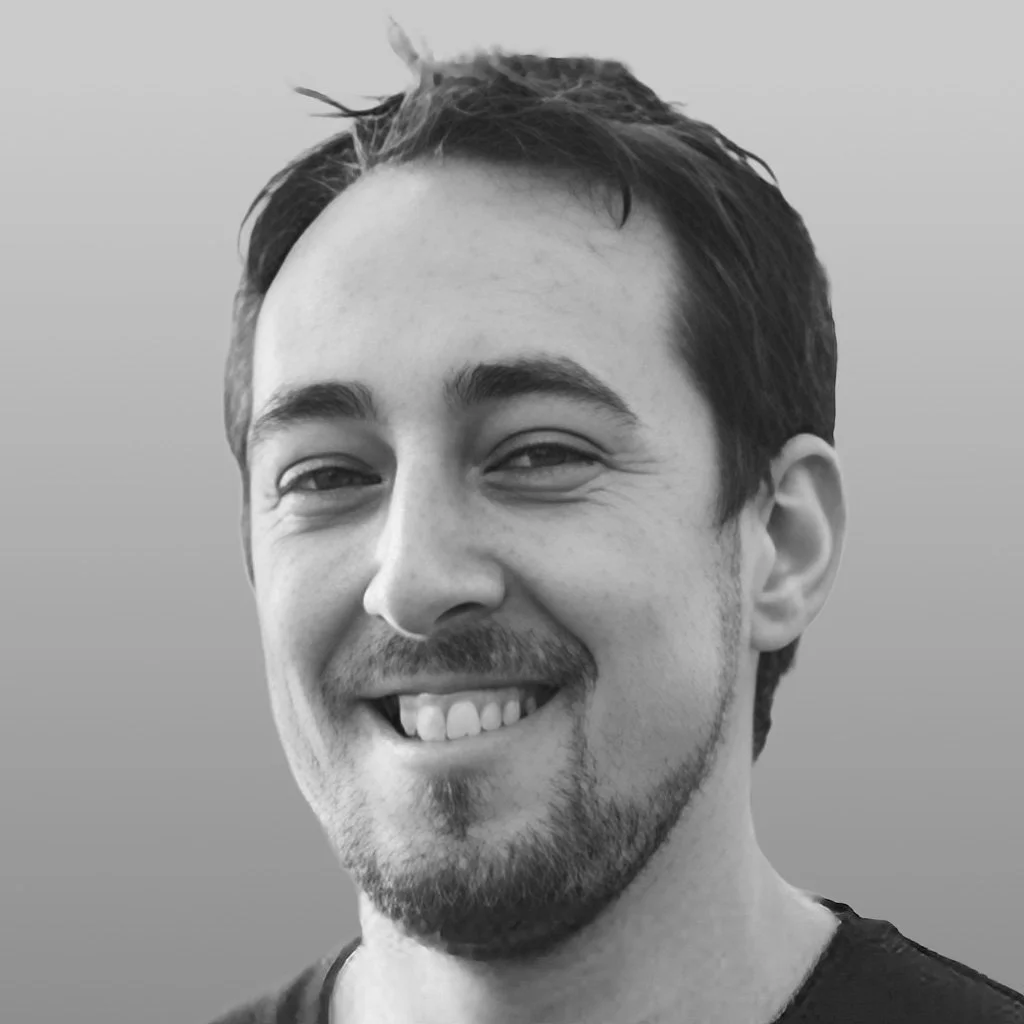 Black and white portrait of a smiling young man with short dark hair and facial hair, wearing a black shirt.