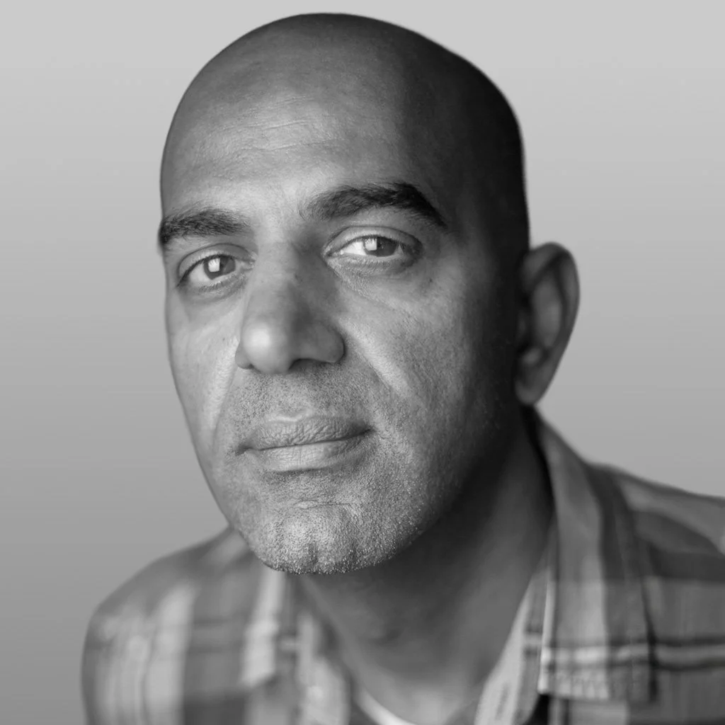 Close-up black and white portrait of a man with a serious expression, short-cropped hair, and wearing a plaid shirt.