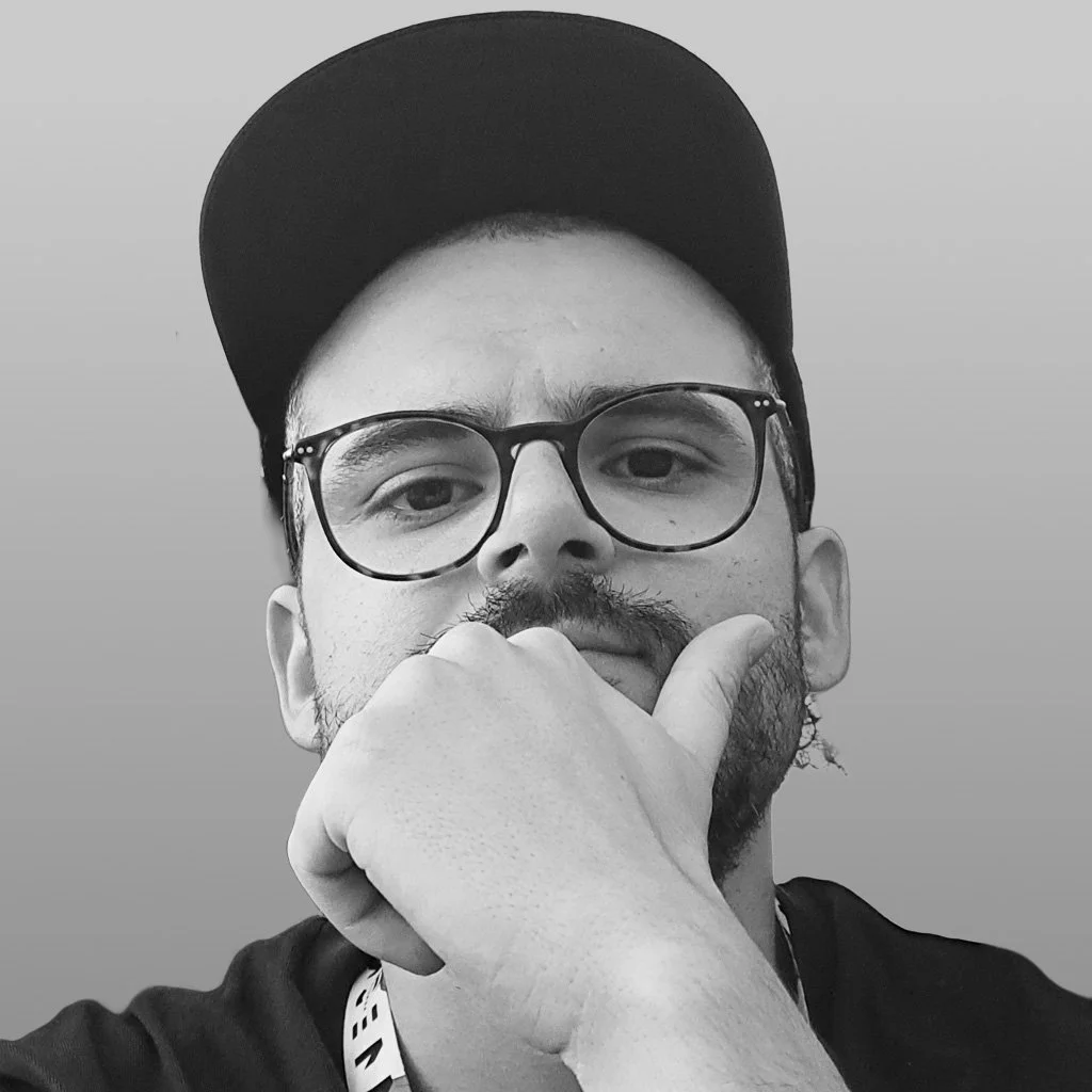 A young man with glasses and a beard, wearing a cap and a black shirt, resting his chin on his hand with a thoughtful expression.