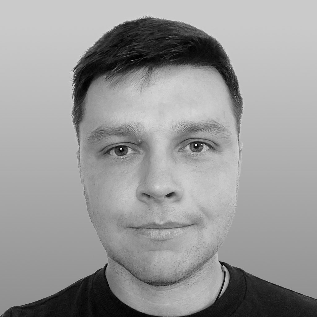 Black and white close-up portrait of a man with short dark hair, light skin, and wearing a dark shirt, looking directly at the camera against a plain background.