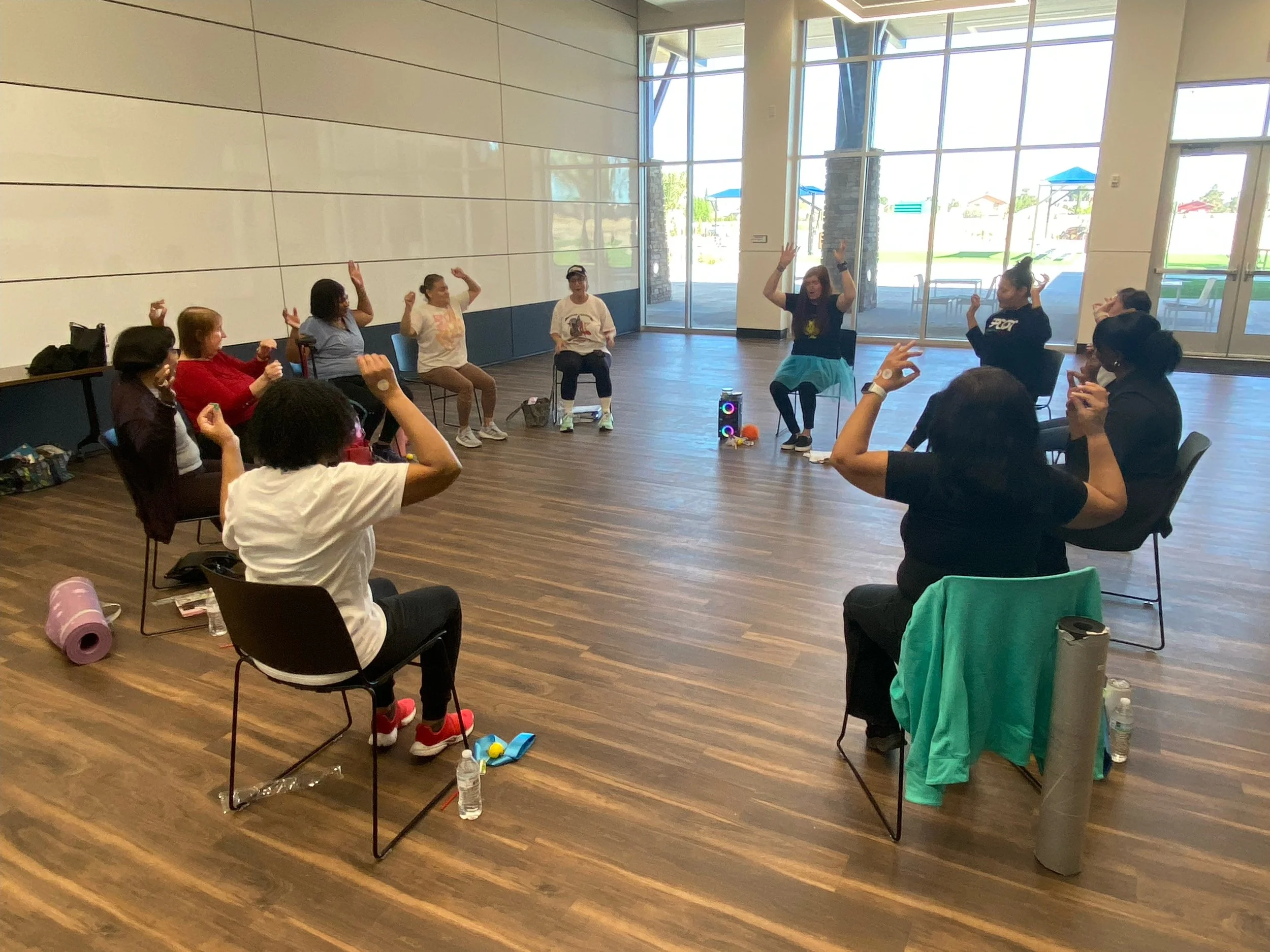 Group of people, mostly women, participating in a seated exercise or dance class in a bright, spacious room with large windows, wooden flooring, and casual attire.