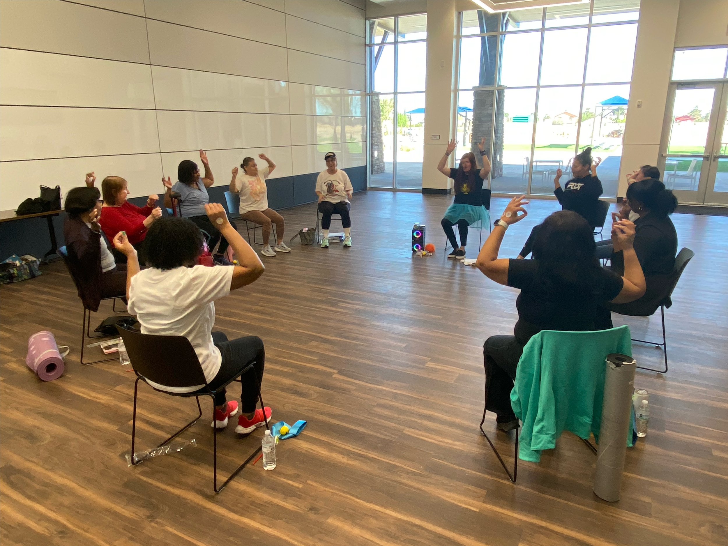Group of women participating in a seated exercise class inside a spacious room with large windows. They are raising their arms and engaging in physical activity, with some smiling and concentrating. Personal belongings and water bottles are nearby.