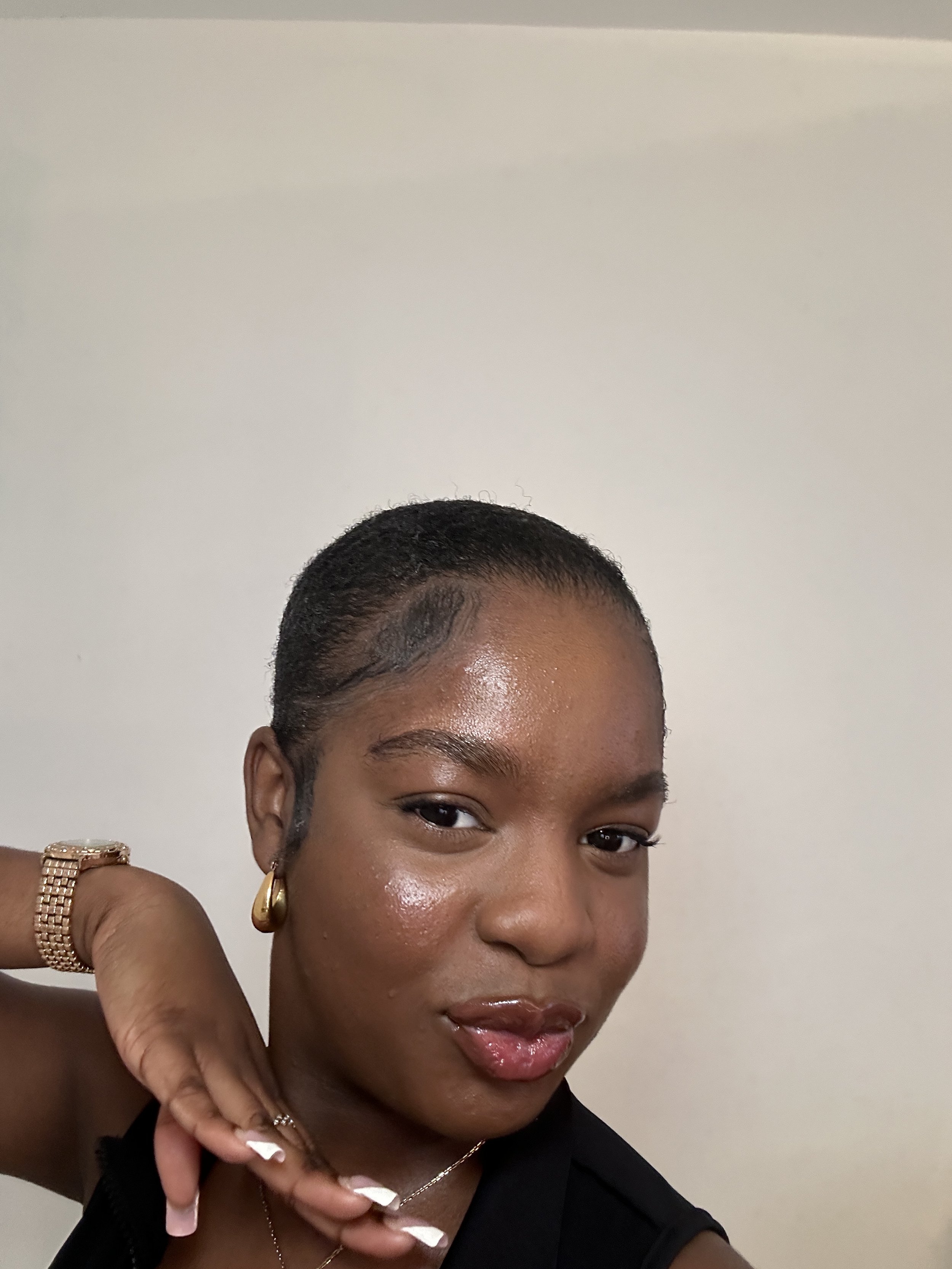 A woman with short, dark hair styled close to the scalp, wearing gold hoop earrings, a gold bracelet, and a ring, posing with her hand near her face against a plain light-colored wall.