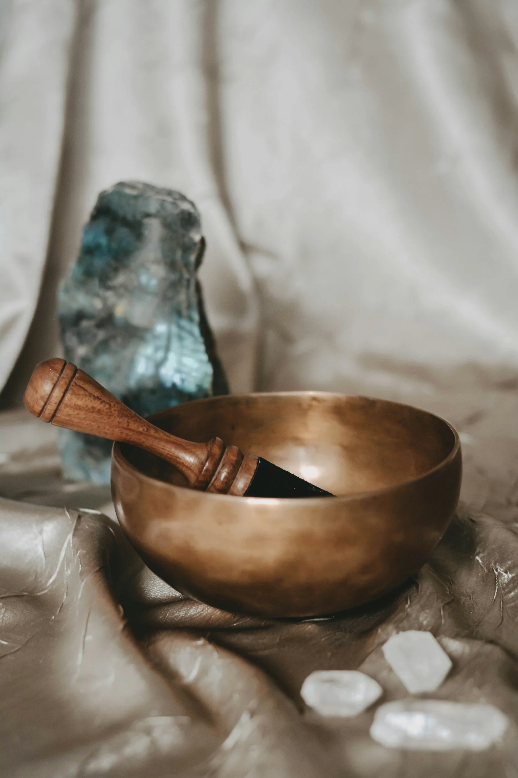 A copper singing bowl with a wooden mallet resting inside, a crystal formation, and some translucent stones on a beige fabric surface.