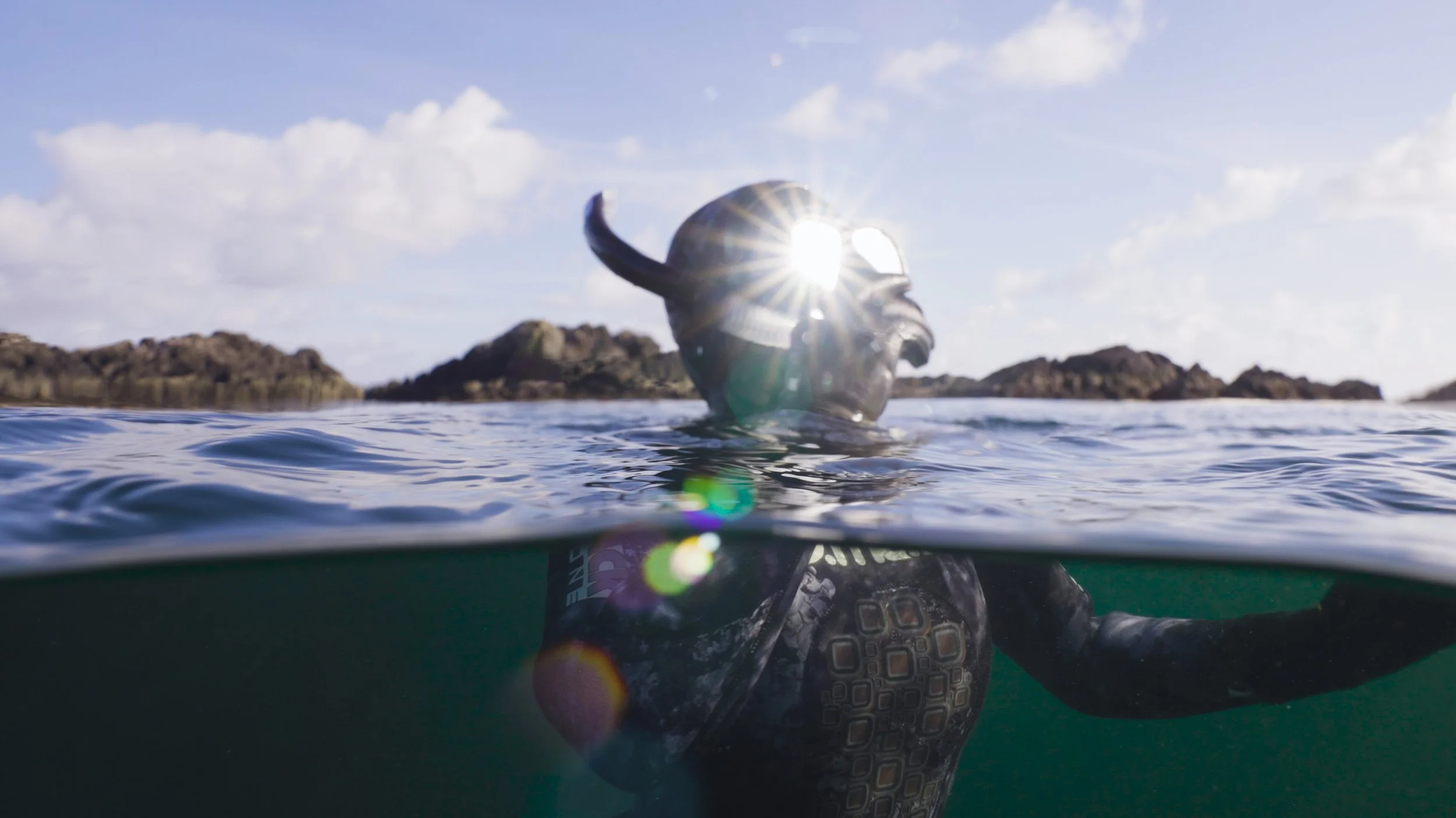 A diver wearing a helmet and wetsuit in the water, partially submerged, with rocky islands and a cloudy sky in the background, the sun creating a flare on the helmet.