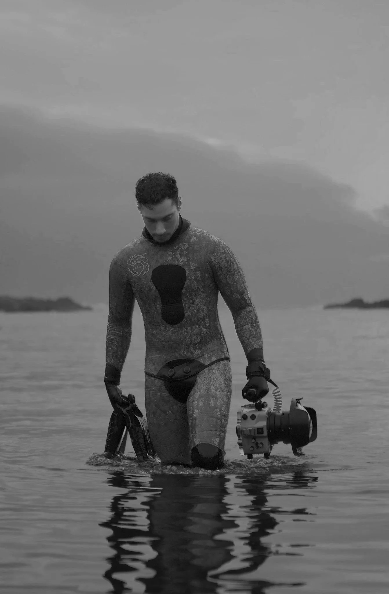 A water cinematographer in a wetsuit standing in the water, holding a professional camera in his left hand, swimming fins in his right, with a cloudy sky in the background.