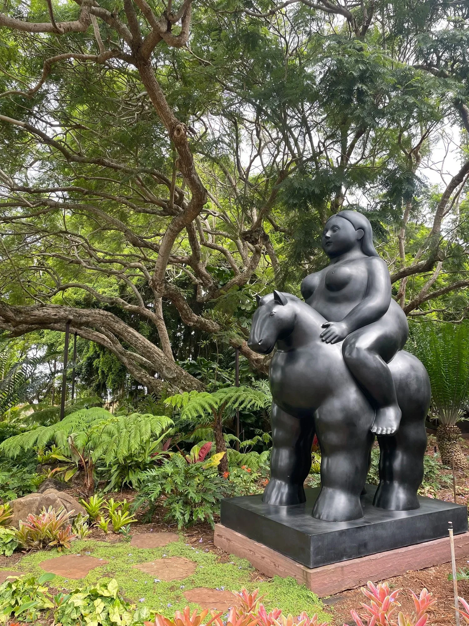 Dark bronze sculpture of a rounded figure on a horse in a garden