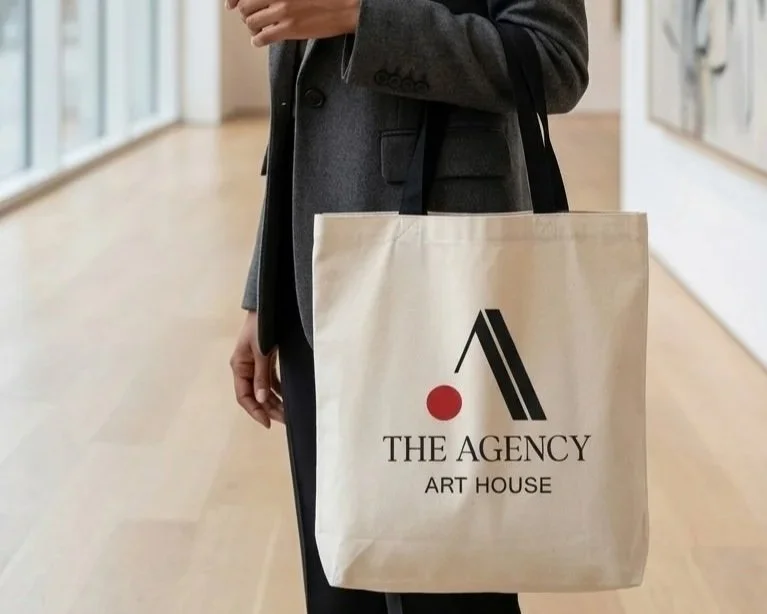 Person standing in an art gallery holding a beige tote bag with "The Agency Art House" logo.