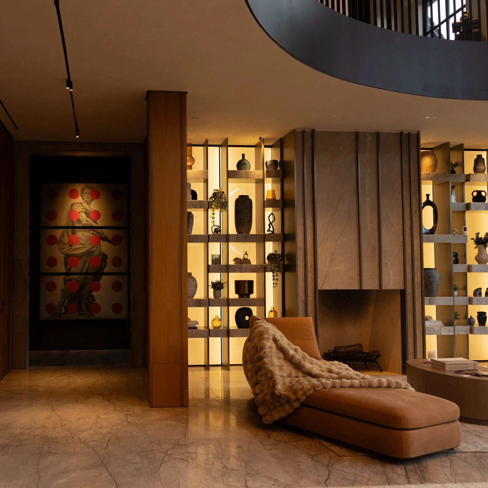 A modern living room in Los Angeles with a tan chaise lounge draped with a plush blanket, a fireplace, and decorative shelving with various vases, sculptures, and plants. Artwork with a classical figure and red dots is visible on the left wall.