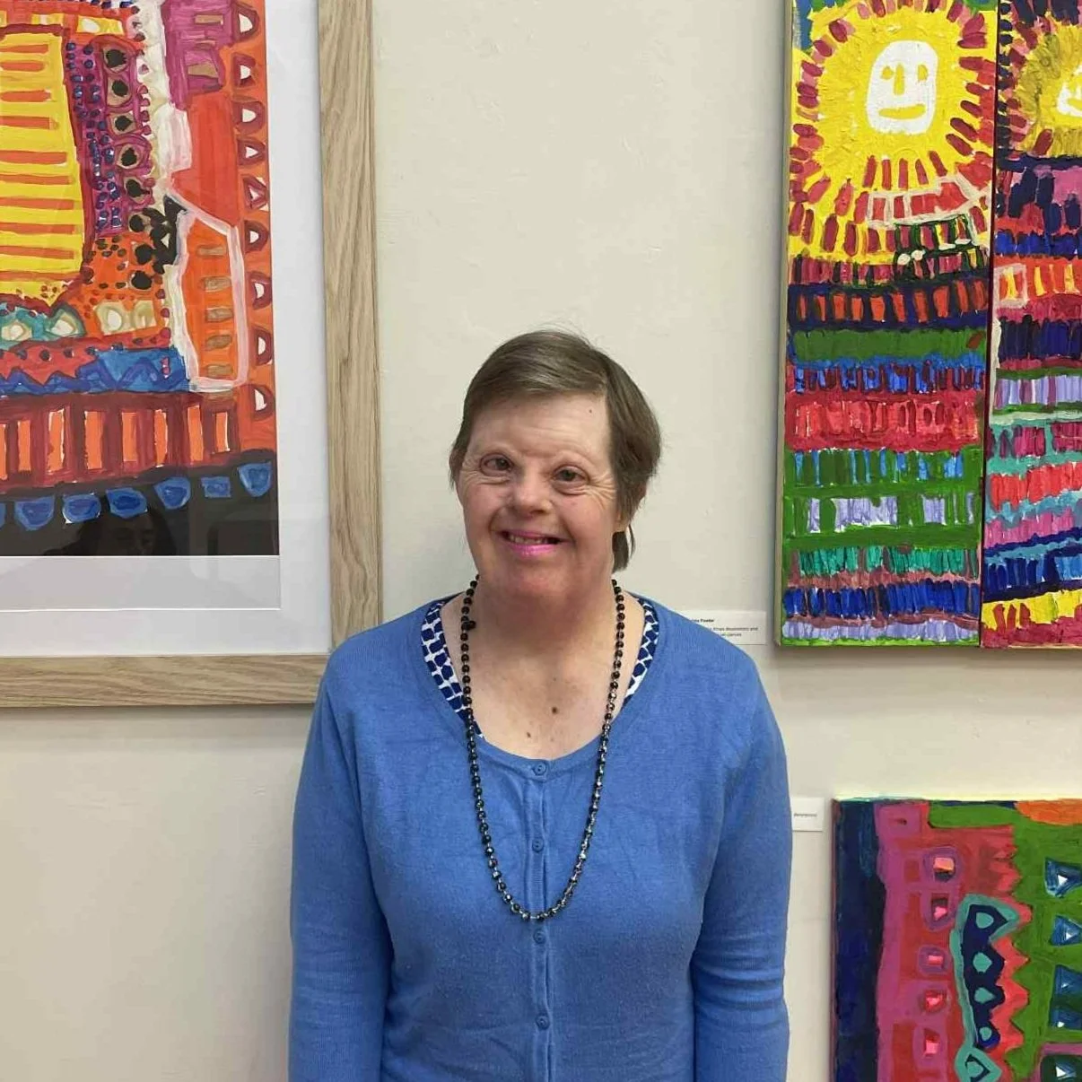 A person standing in front of colourful art hung on a cream wall. The person is in a blue cardigan. The art features bright patterns in multiple colours.