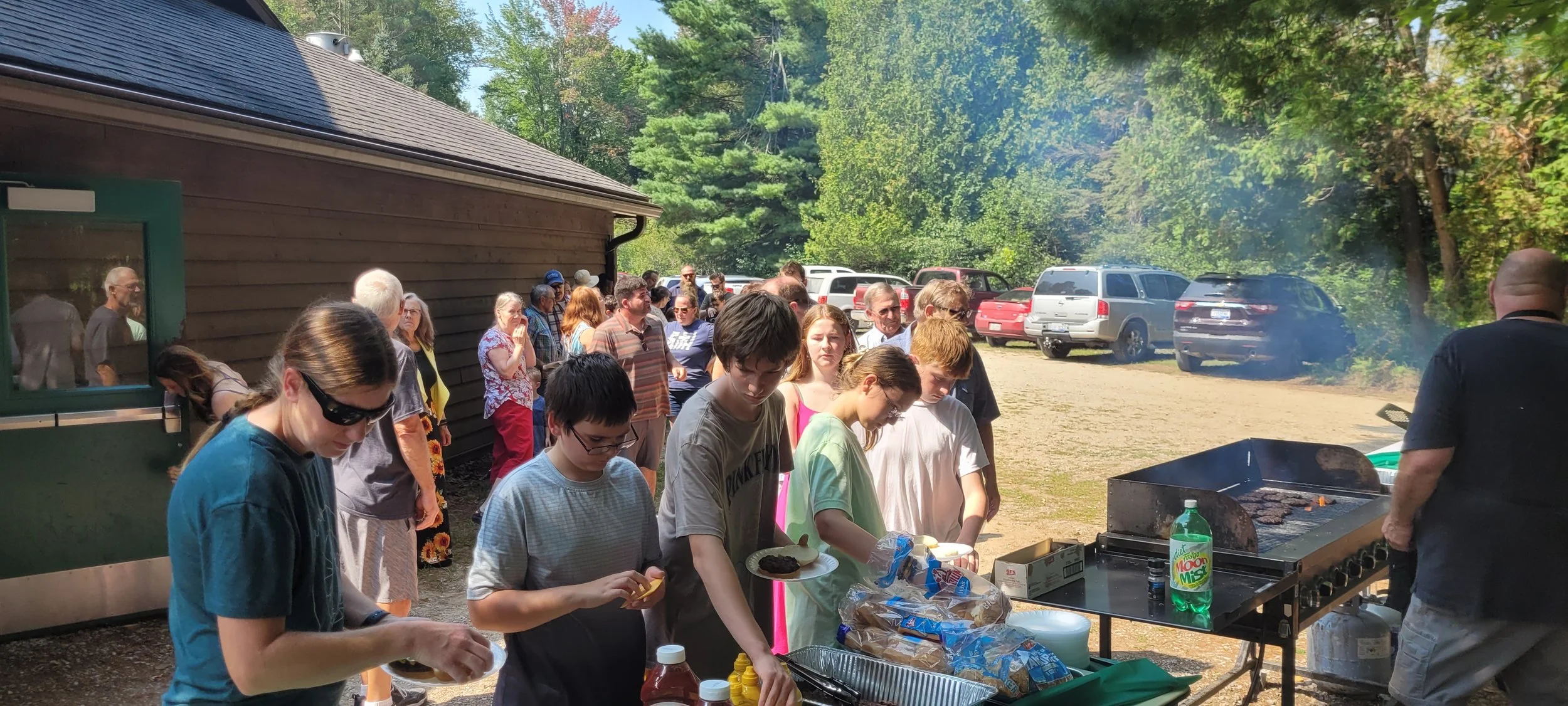 A group of people standing in line at a picnic barbecue outdoors surrounded by trees and parked cars, with a griddle cooking meat and people preparing food.