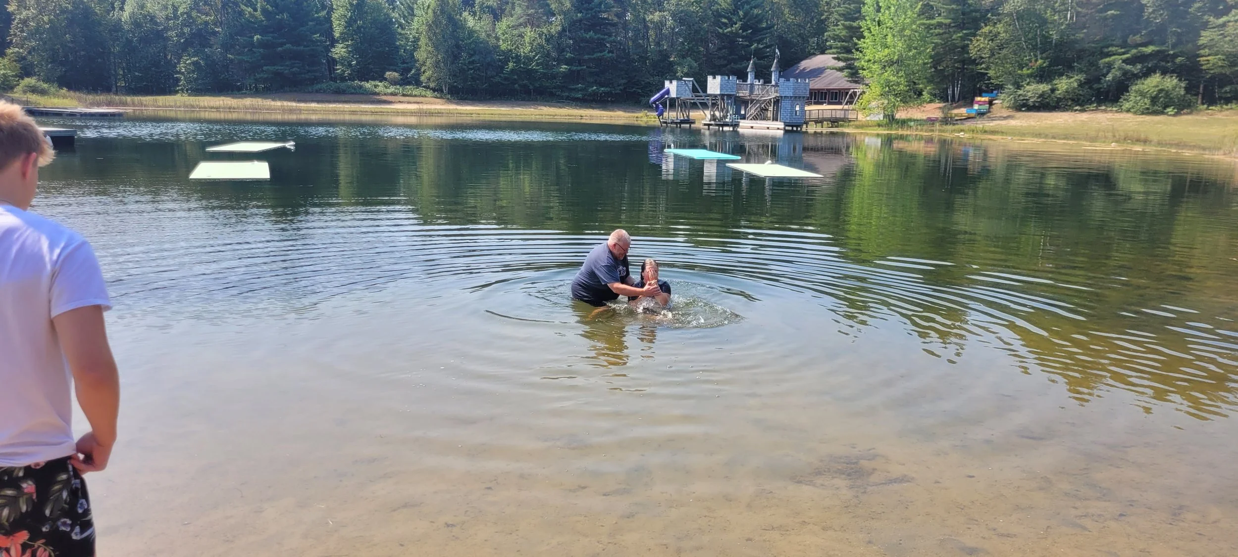 A man is baptizing a person in a lake, with a boy in a white shirt and shorts watching nearby. In the background, there is a castle-like structure on the shore and a forest.
