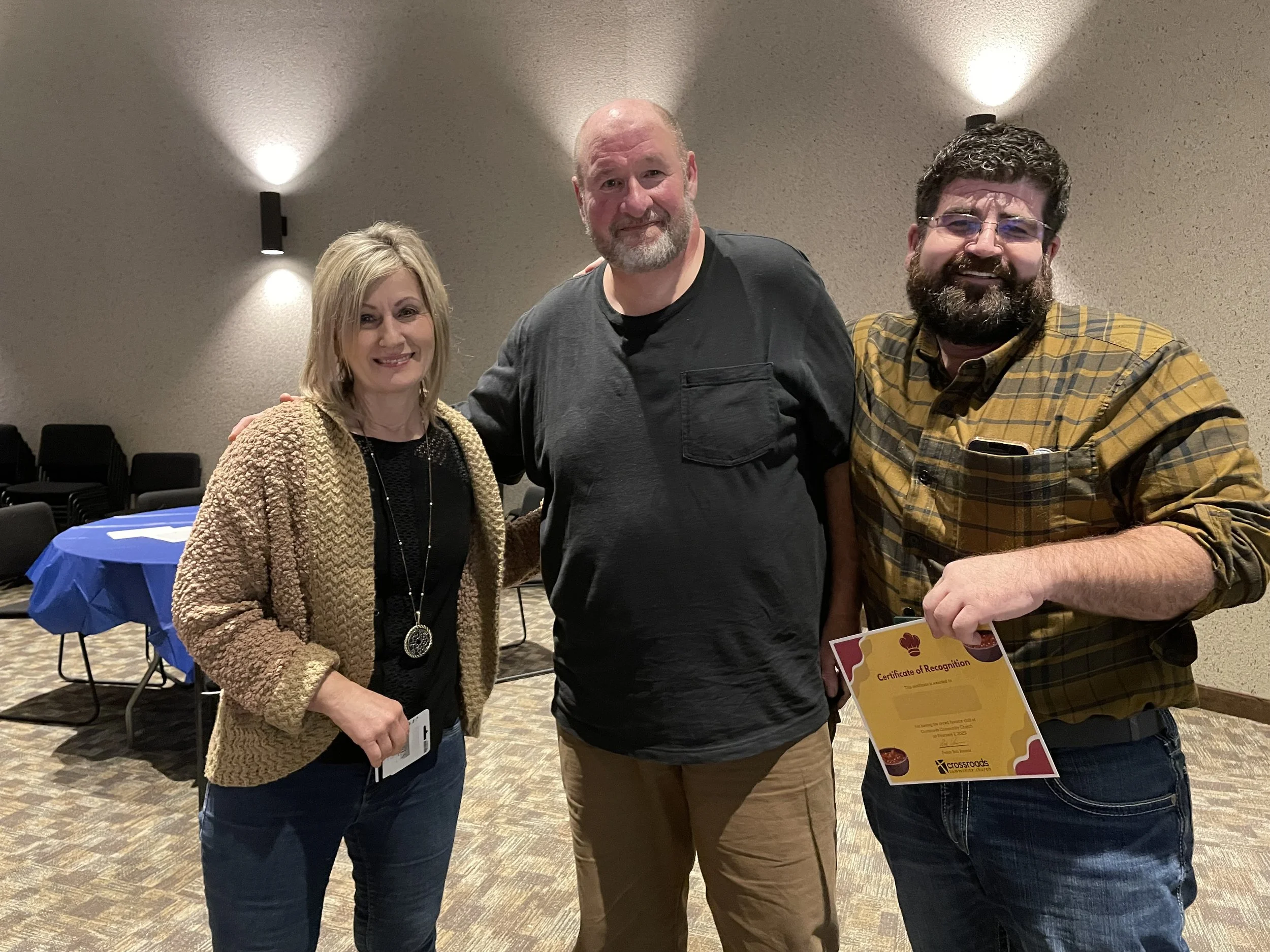 Three people standing together in an indoor setting, with two holding certificates, smiling at the camera.