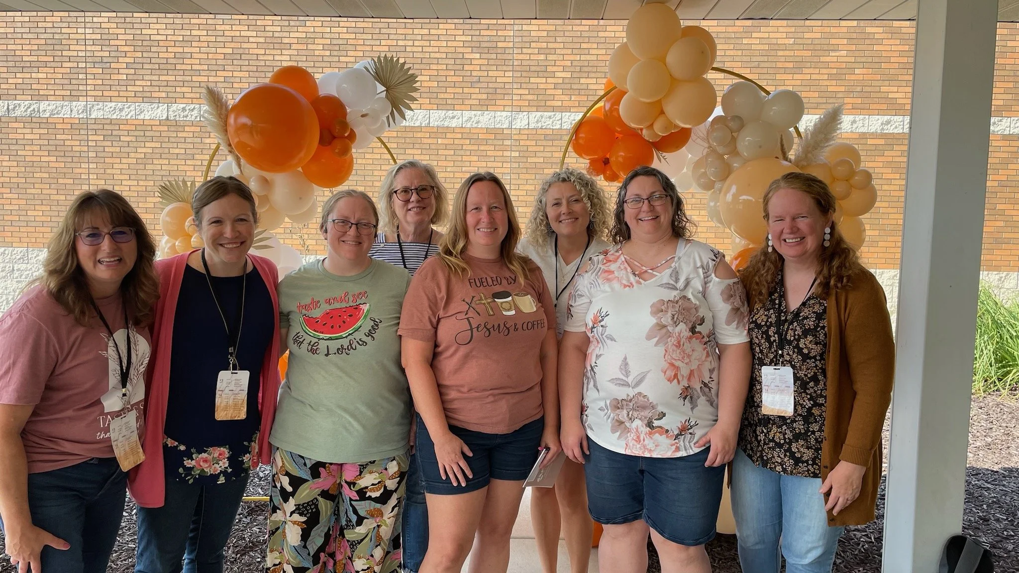 Group of nine women smiling, standing under a decorative balloon arch with orange, white, and beige balloons, in front of a brick wall, at an outdoor event or gathering.