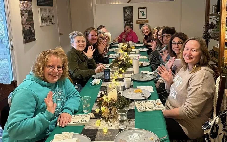 A group of women sitting around a long dining table, smiling and waving at the camera during a gathering or celebration.