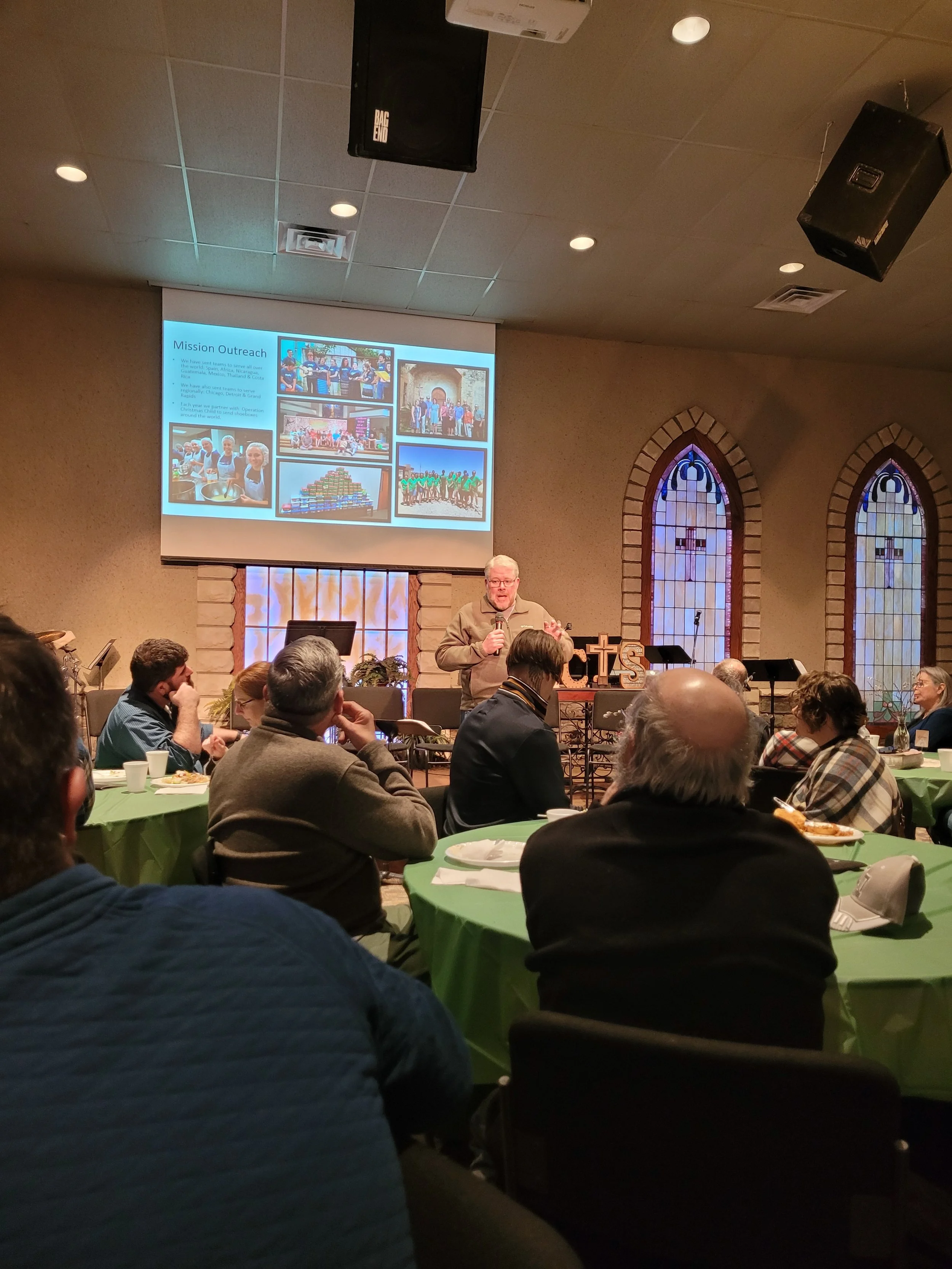 A man giving a presentation in a church setting. Several people are seated at round tables covered with green tablecloths, listening. A large screen behind the speaker displays a presentation slide titled "Mission Outreach" with images of community a