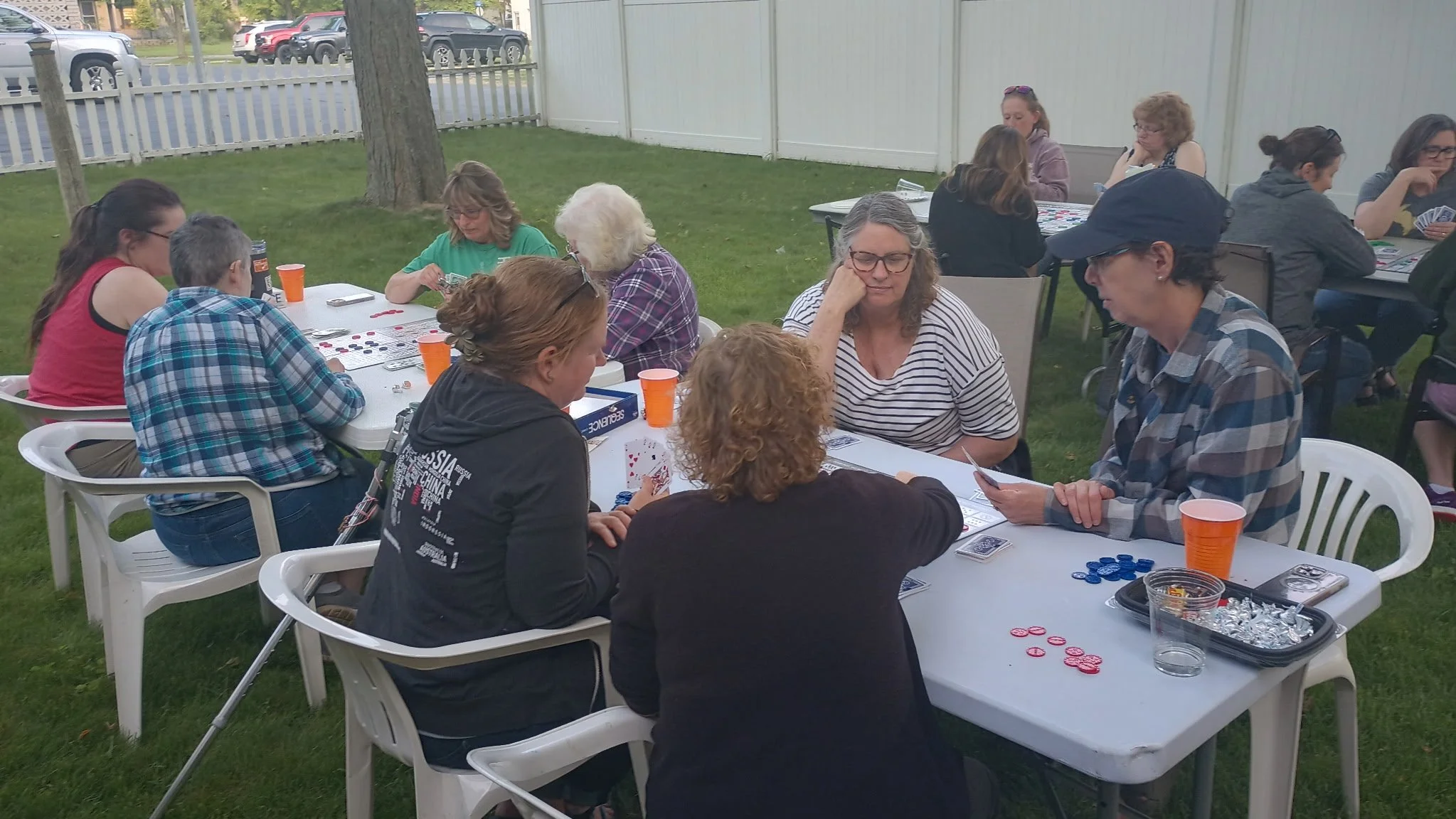 Group of people playing board games outdoors at a garden party, sitting around white plastic tables with orange cups, with trees, a wooden fence, and parked cars in the background.