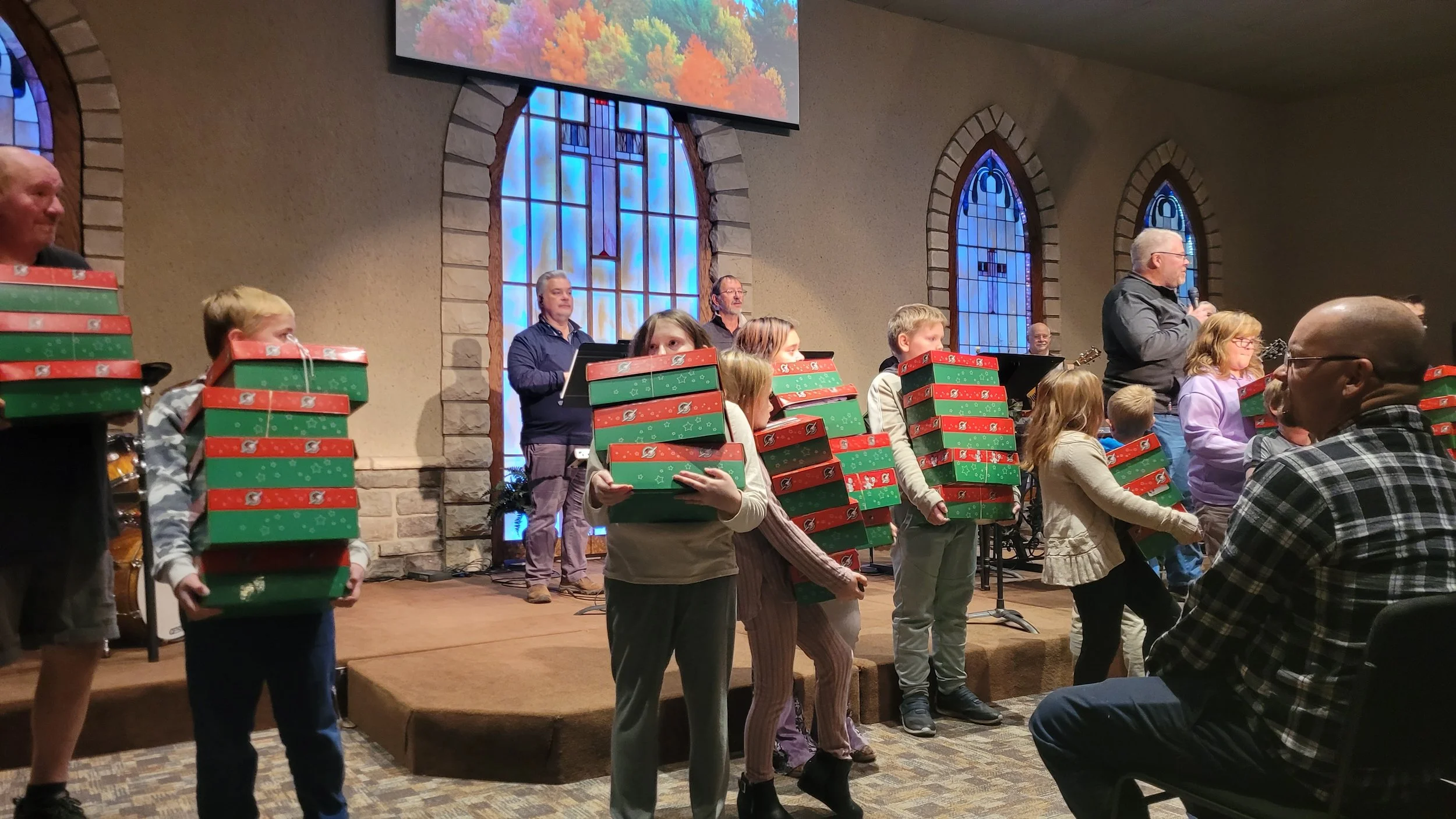Children holding stacked Christmas gift boxes on stage during a holiday event in a church, with adults and a band in the background and stained glass windows.