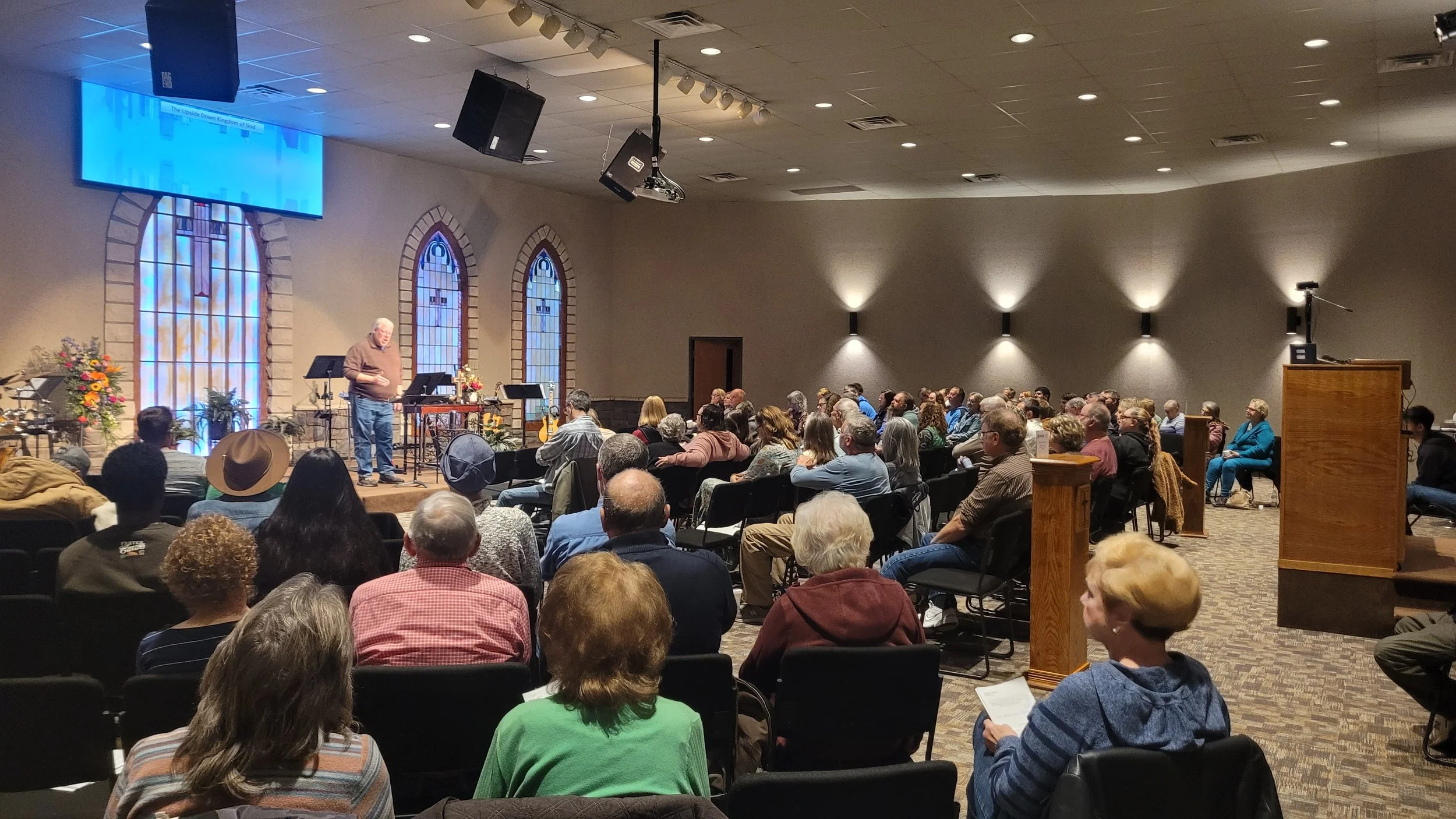 A man speaks to an audience in a church or conference hall with stained glass windows, floral arrangements, and a large screen overhead. People are seated facing the speaker, listening attentively.