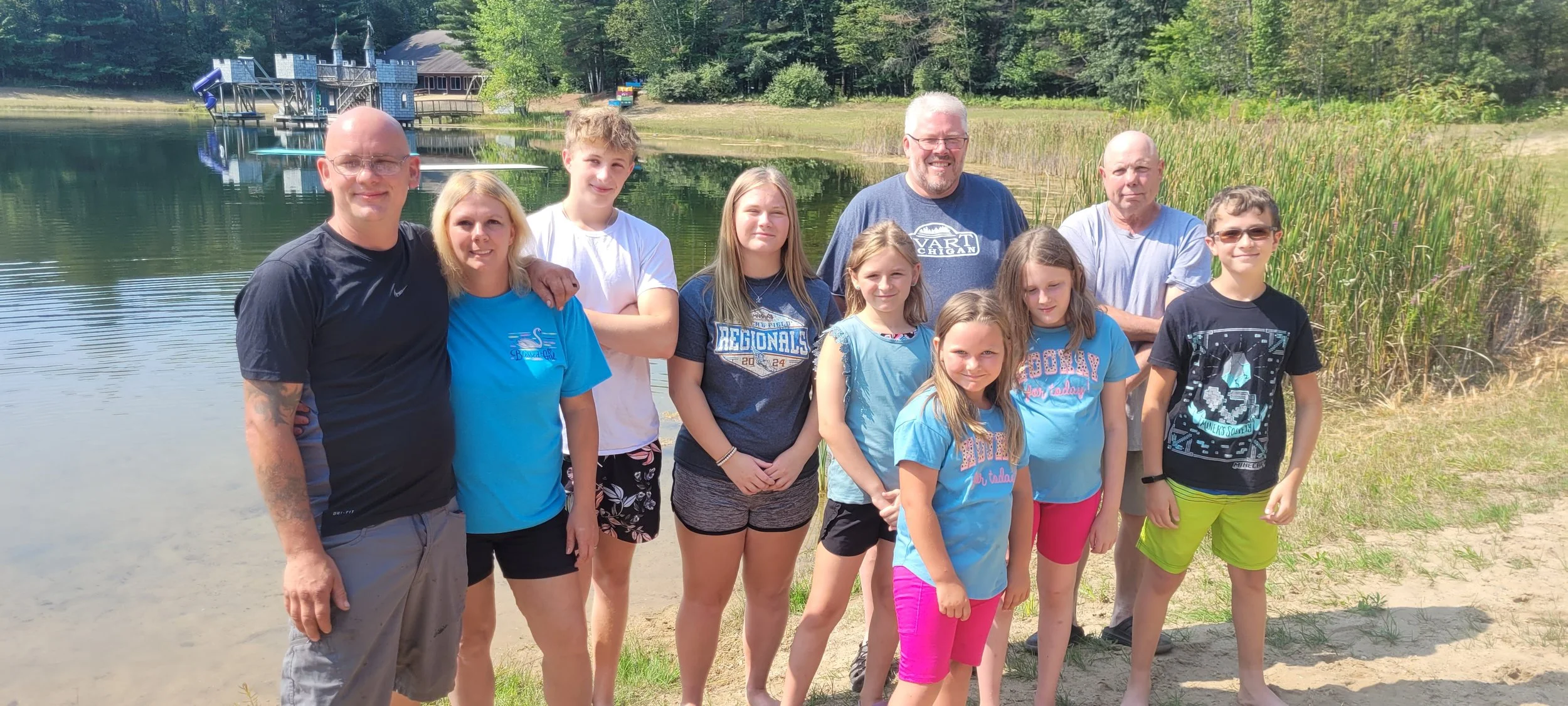 Family or group of people standing on a sandy area by the lake, smiling, with trees and a blue play structure in the background on a sunny day.