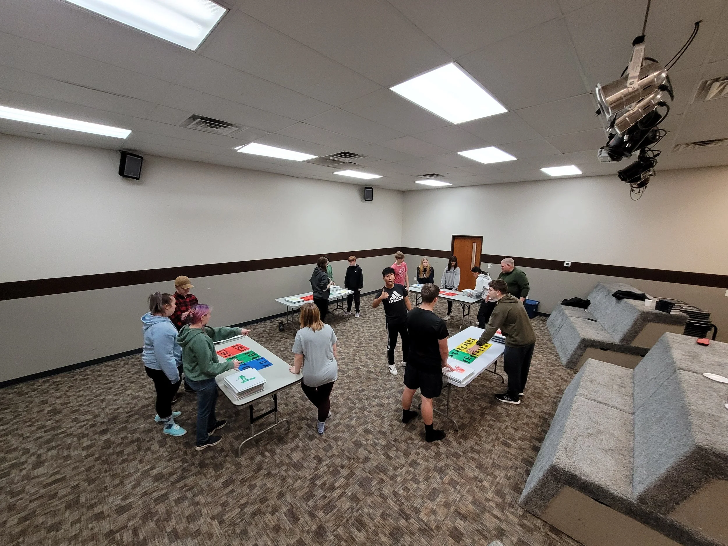 Group of people gathered around tables with colorful signs and papers in a room with beige walls and a brown carpet, preparing for an activity or workshop.