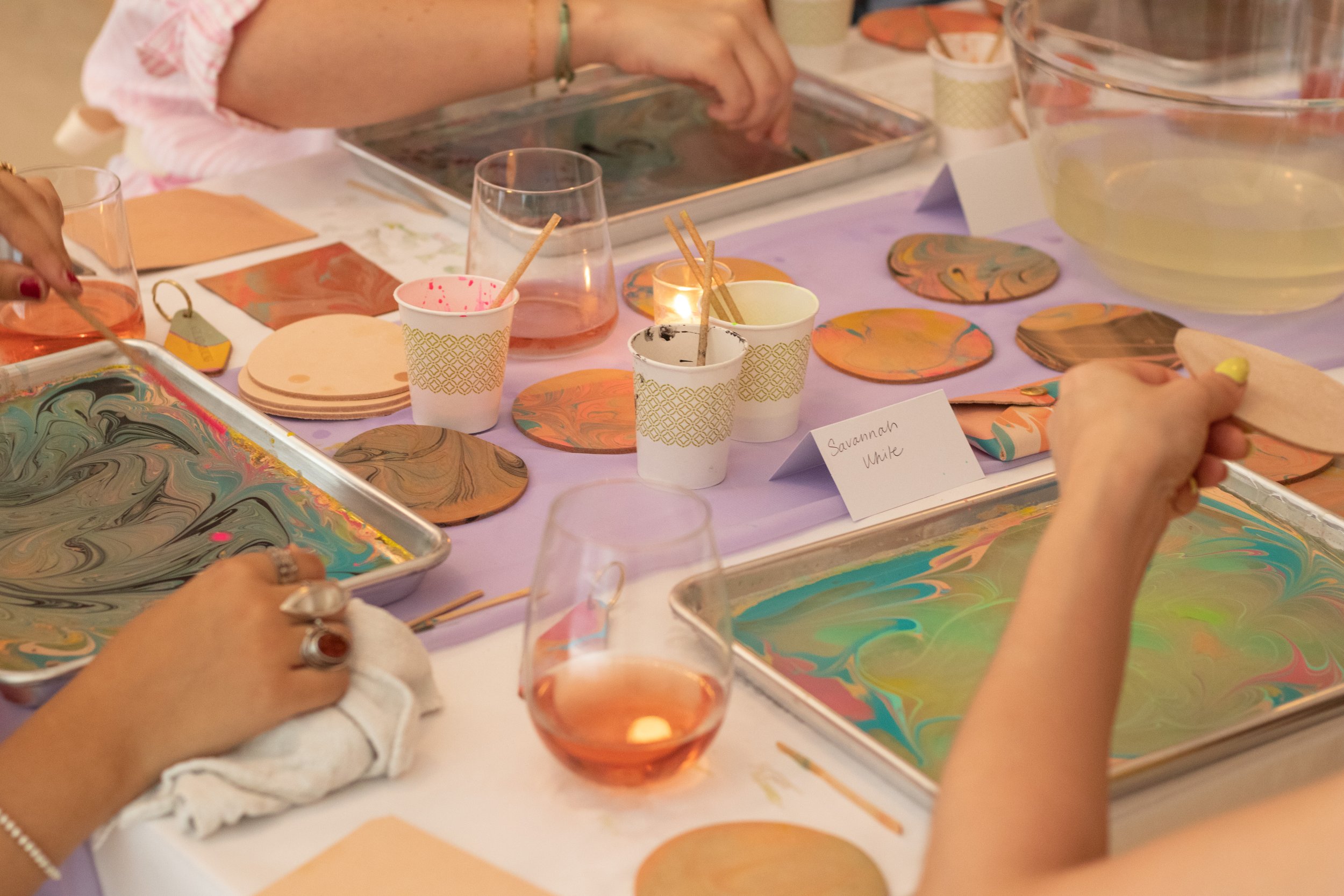 People doing marbling art with colorful paints and circular paper, set on a table with cups and trays.