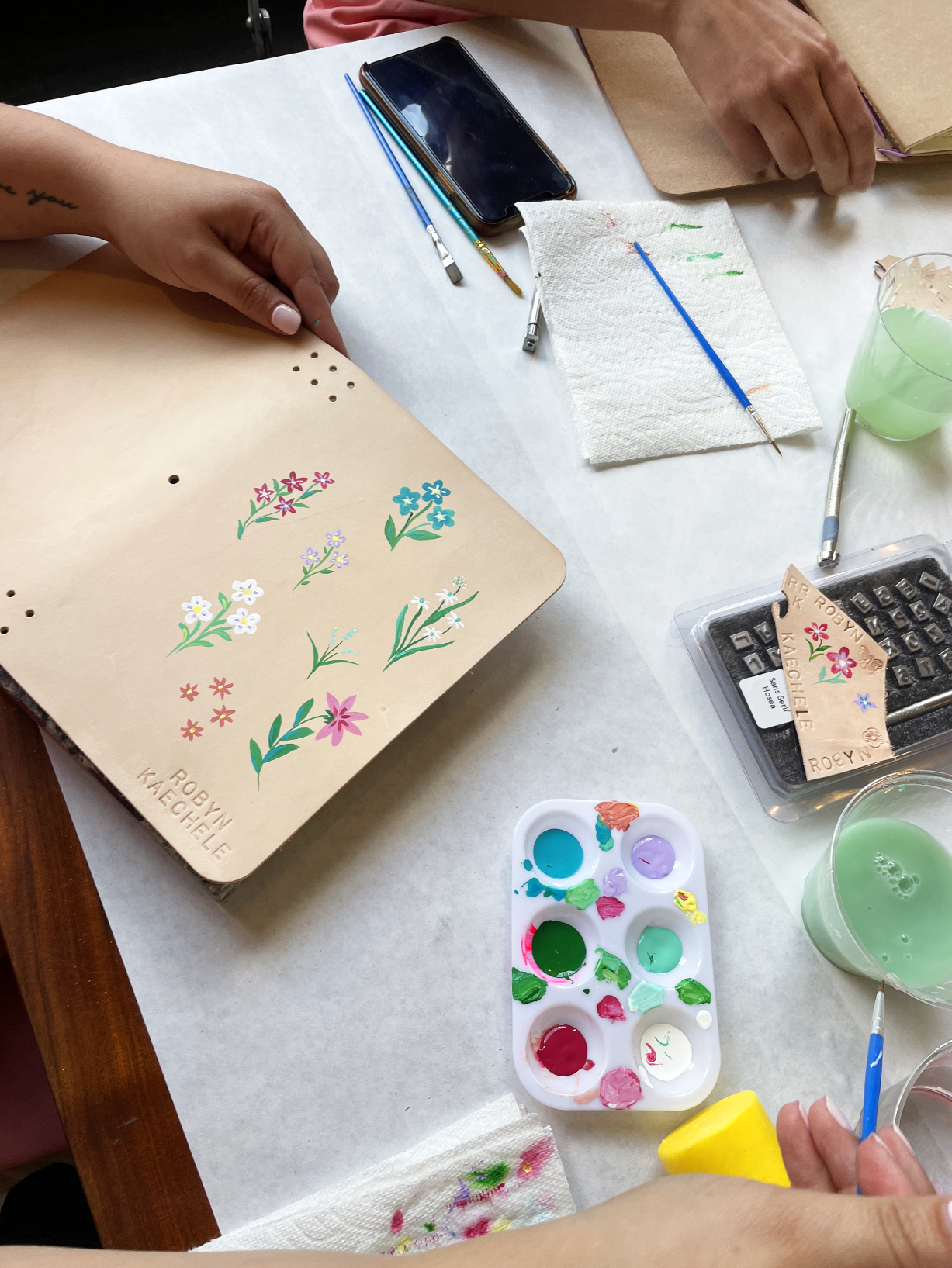 People painting floral designs on a leather item, with brushes, palette, and water cup on a table.