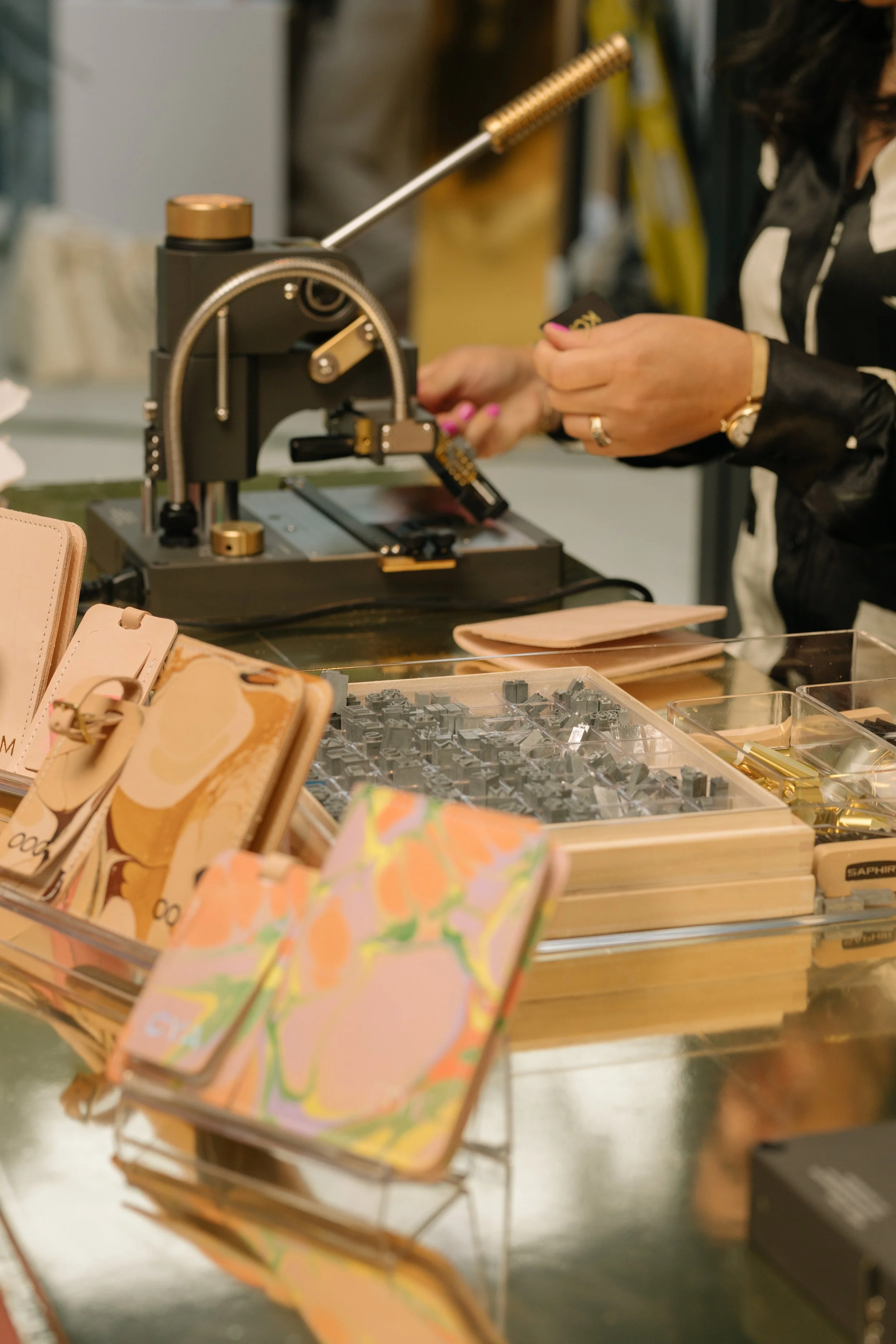 Close-up of a person using a monogram embossing machine with assorted leather goods and stamping tools on a table.
