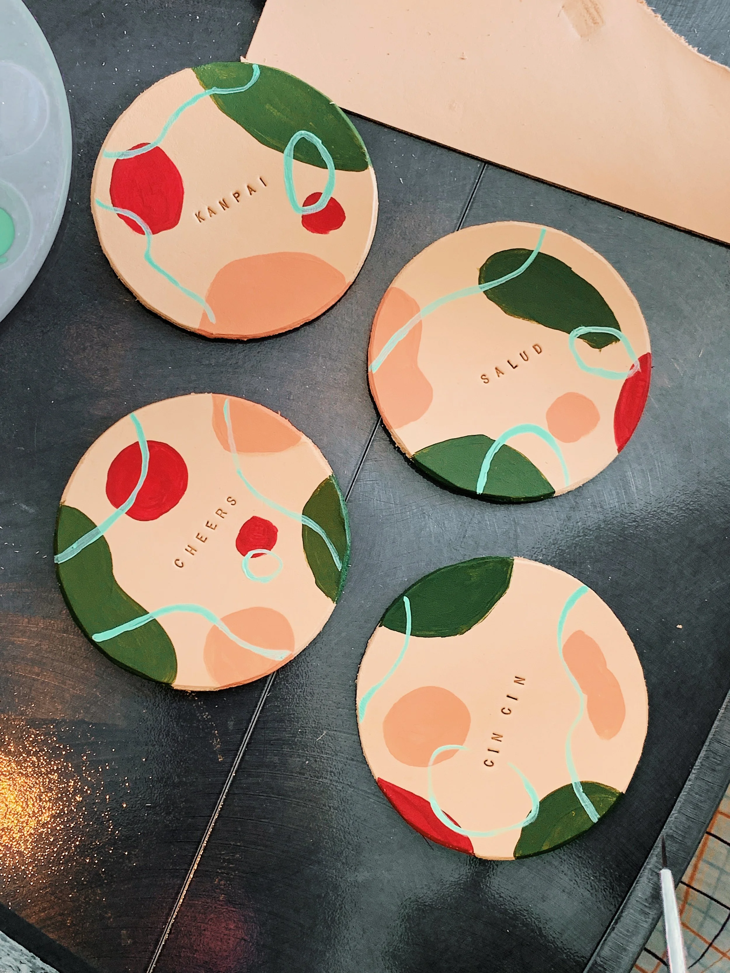 Four round coasters with abstract designs and words "KANPAI," "SALUD," "CHEERS," and "CIN CIN" on a black surface.