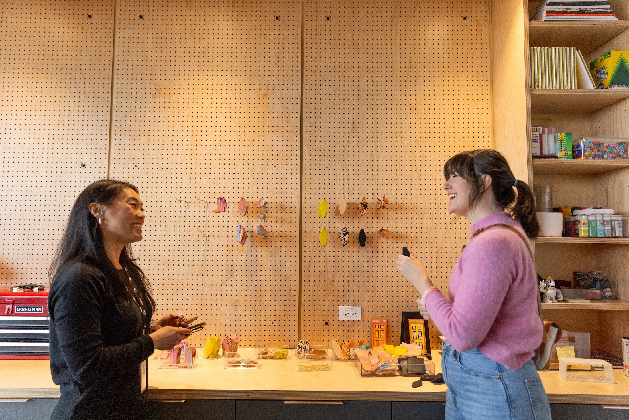 Two women smiling and talking in a workshop with a pegboard wall, crafting materials, and shelves.
