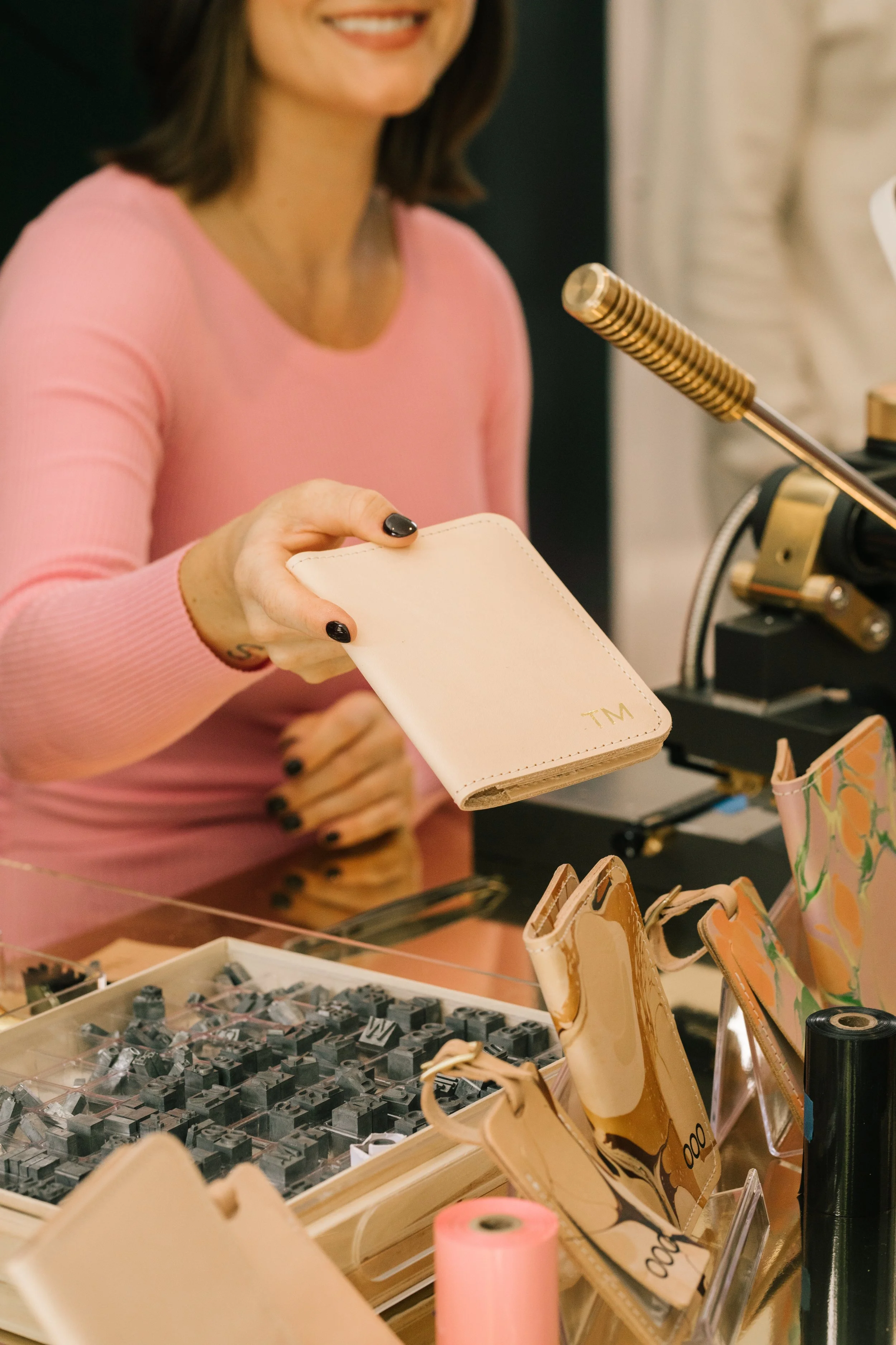 Woman in a pink shirt holding a beige leather wallet with embossed initials, standing near a stamping machine and assorted leather goods.
