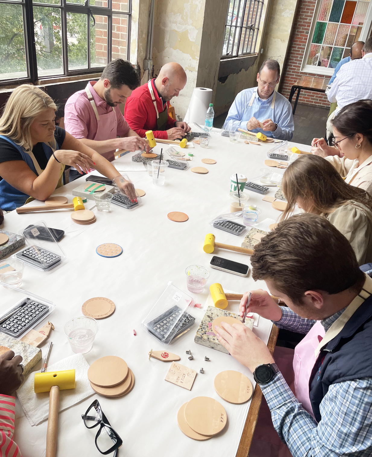 Group of people crafting with leather and stamps around a table, using tools like hammers and cutting mats.
