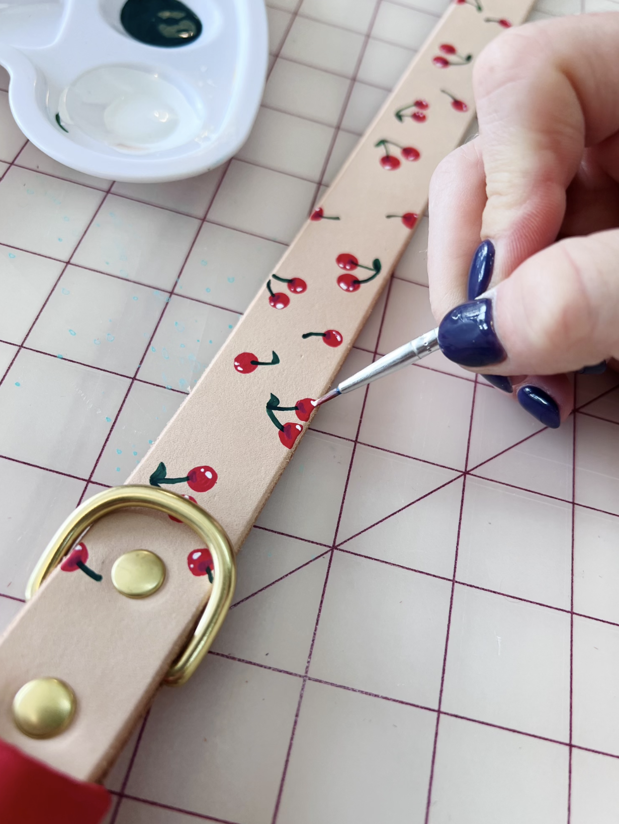 Person painting a leather dog collar with cherry designs, using a small brush. Palette with green paint visible. 