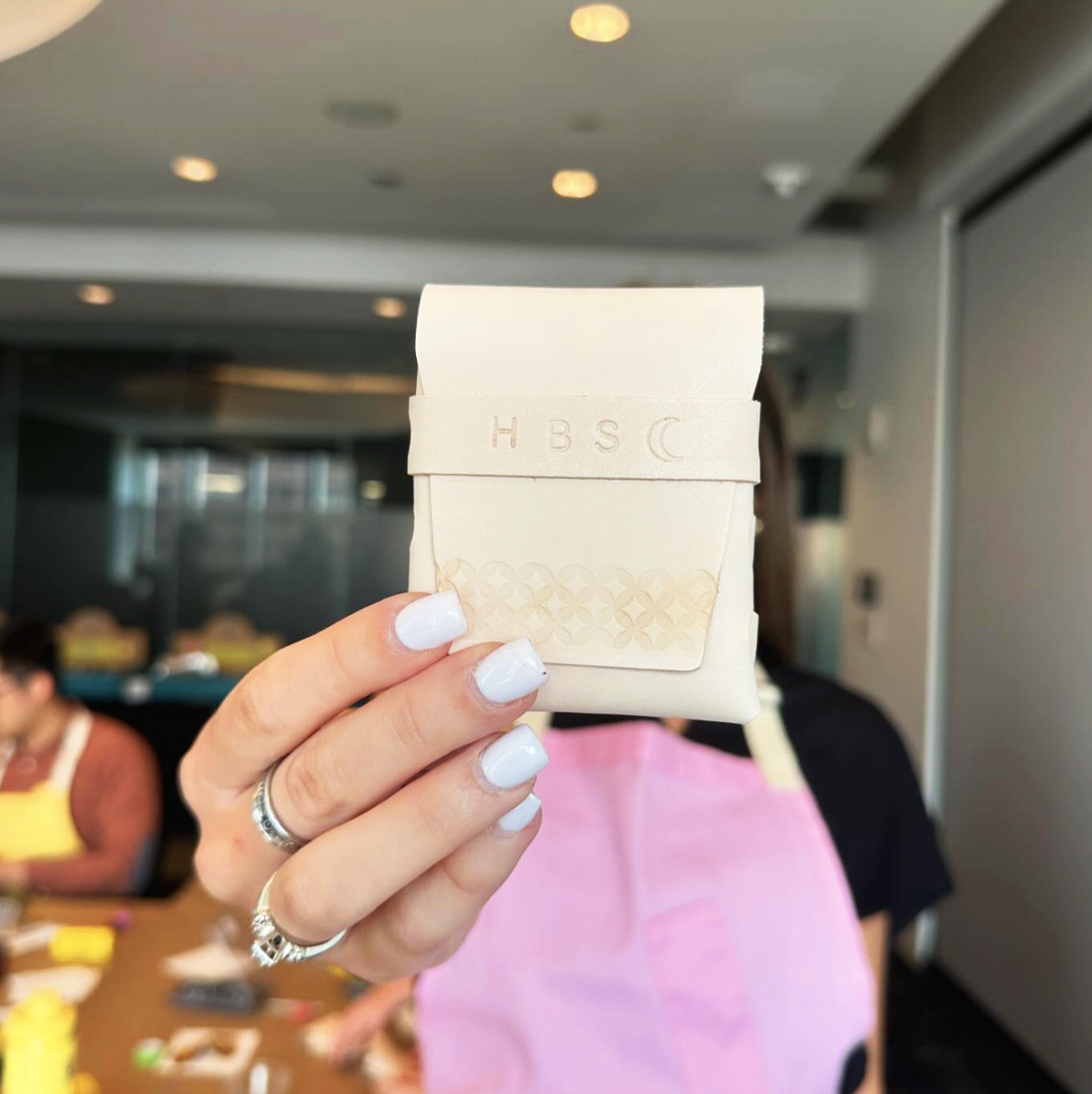 Person holding a small beige leather pouch with embossed letters "HBS" and a crescent moon symbol, in a room with blurred background.