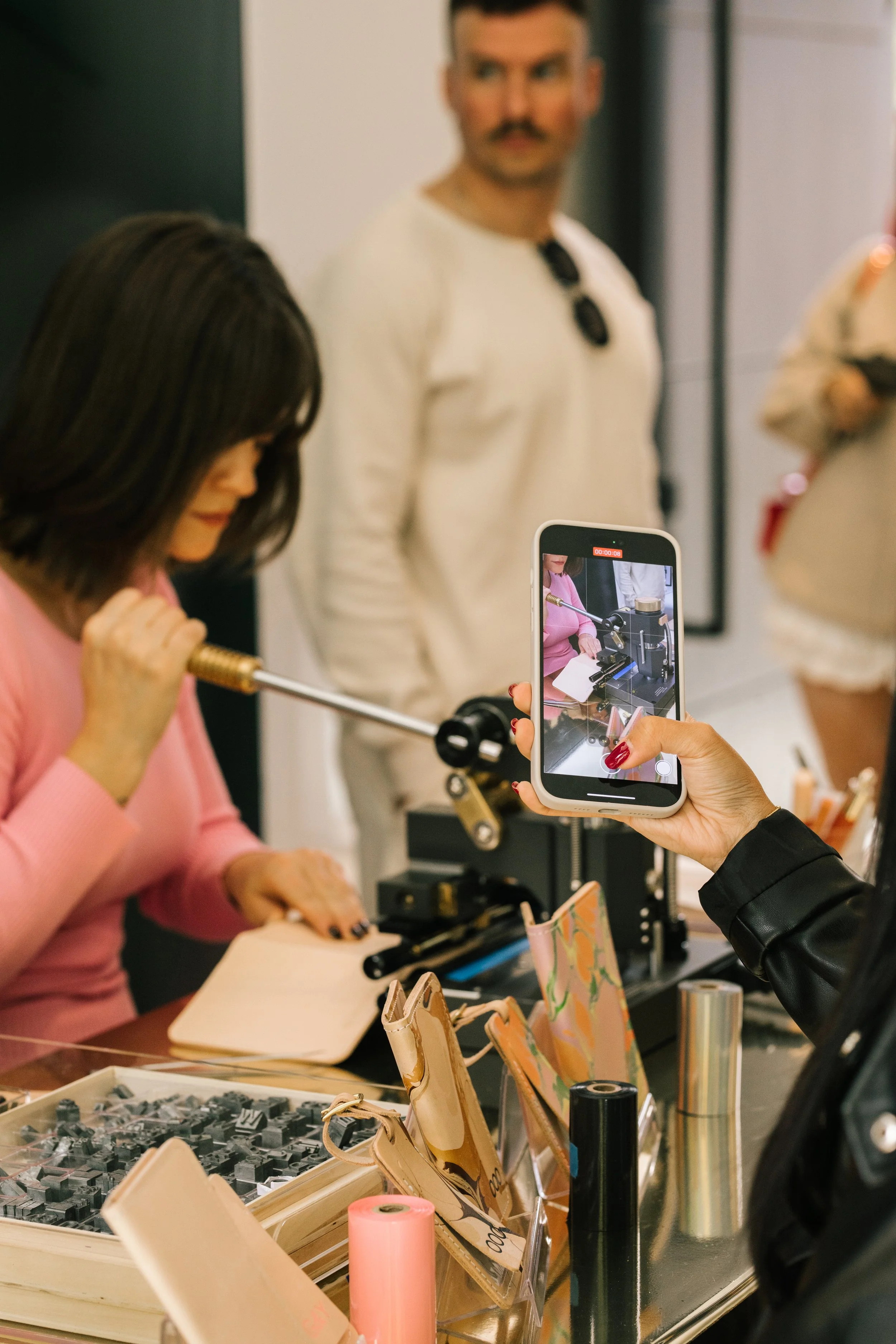 A person in a pink sweater is using a machine to imprint or emboss on a leather item while another person records the process on a smartphone. Various leather goods and tools are on a table.