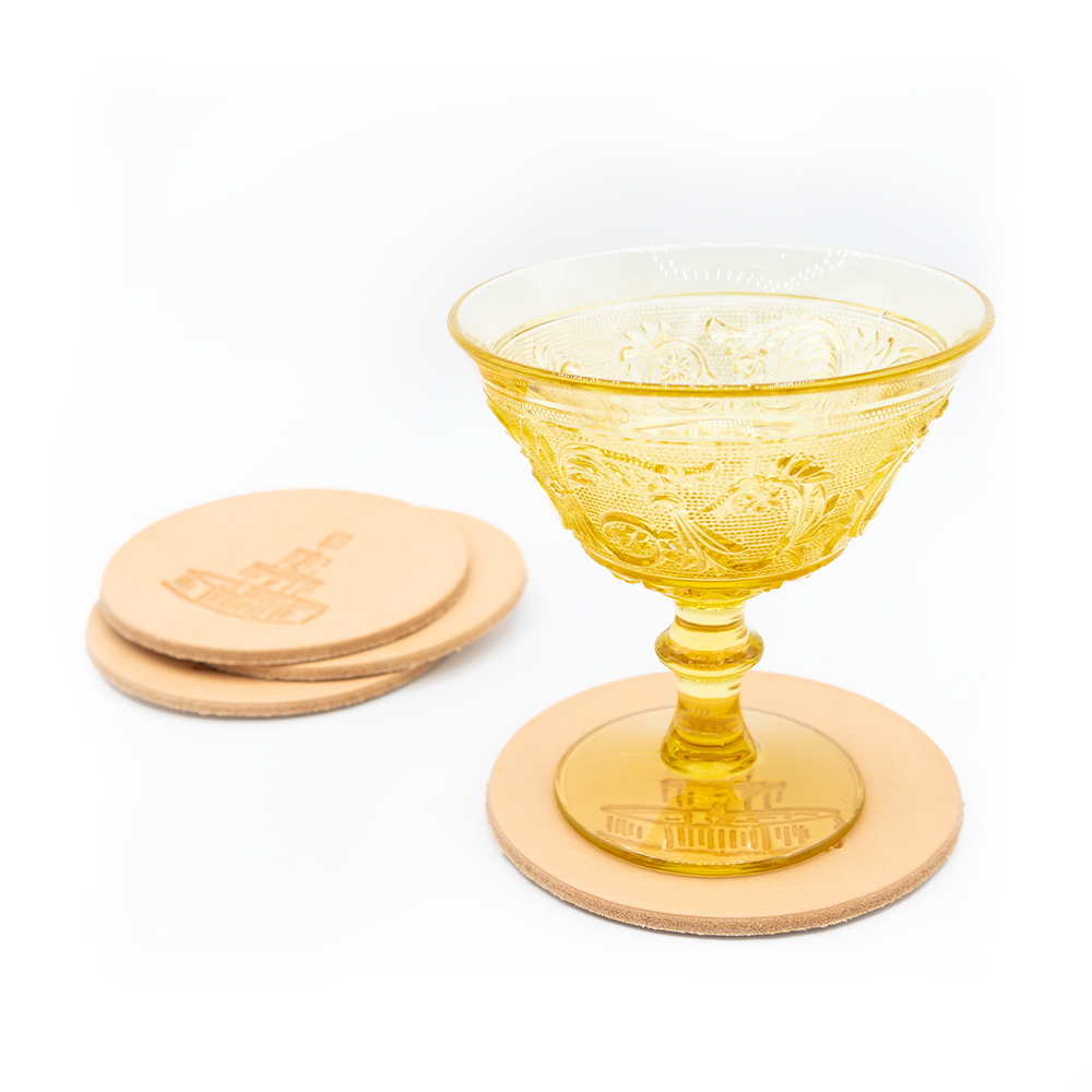 A vintage yellow glass goblet with intricate patterns sits on a round leather coaster. Nearby, there are additional similar coasters with embossed designs.