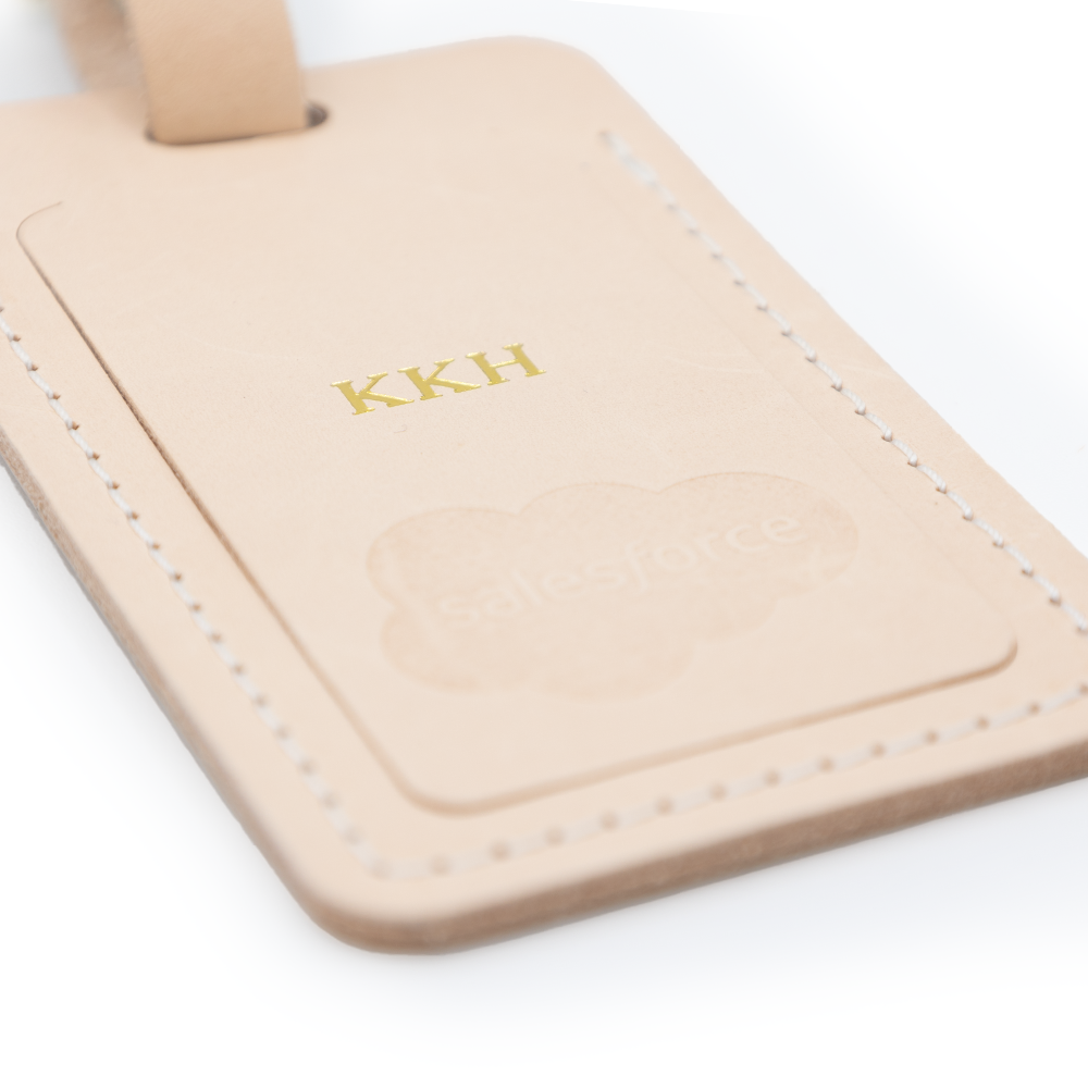 Beige leather luggage tag with "KKH" embossed in gold, "salesforce" logo underneath. Close-up view.