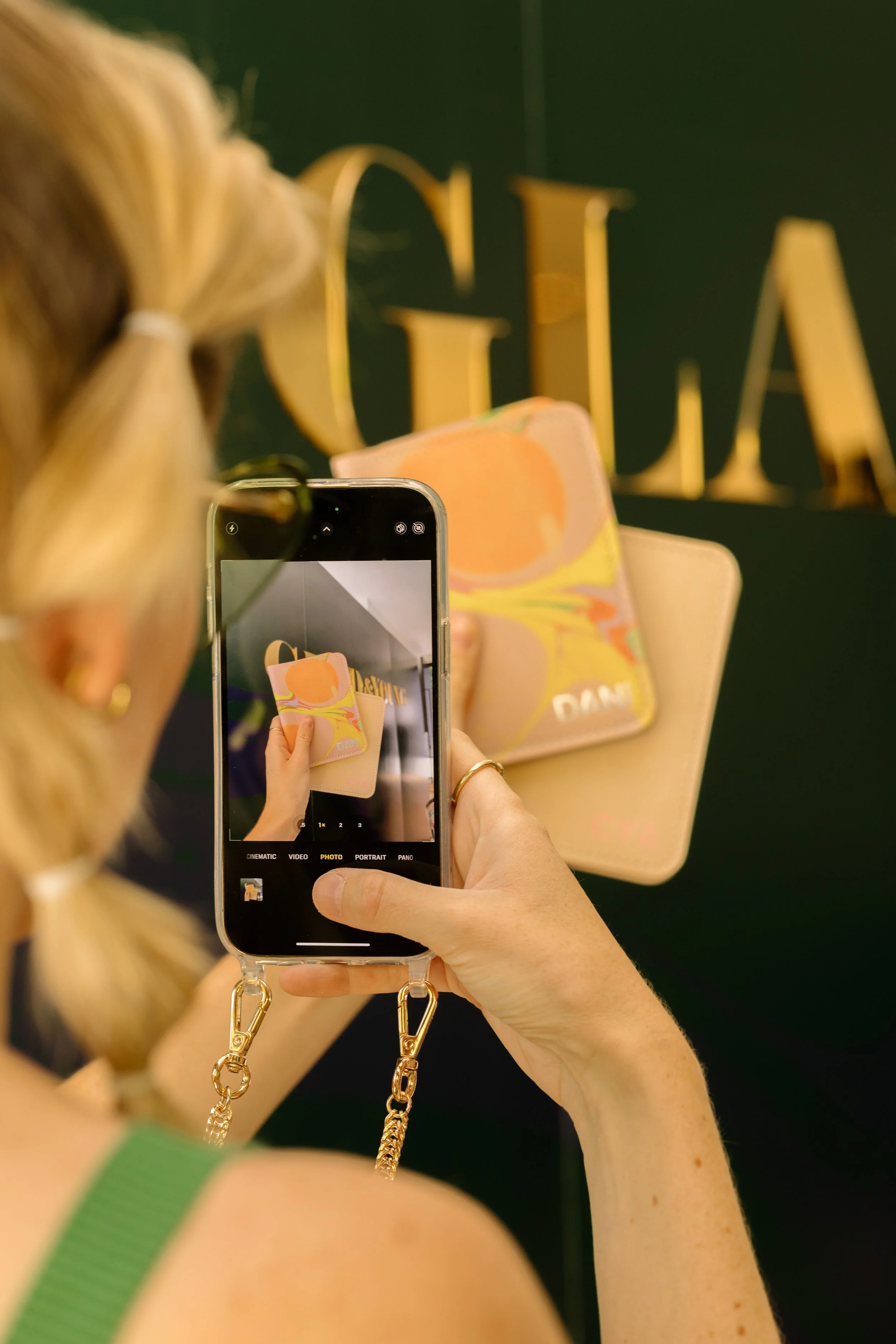 Person photographing a wallet with a smartphone in front of a green and gold background.